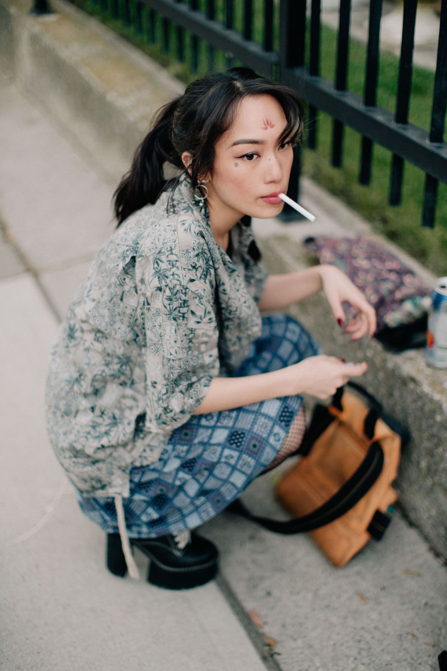 Young Asian woman crouching on sidewalk with cigarette, wearing floral blouse and patterned pants, tan backpack nearby.