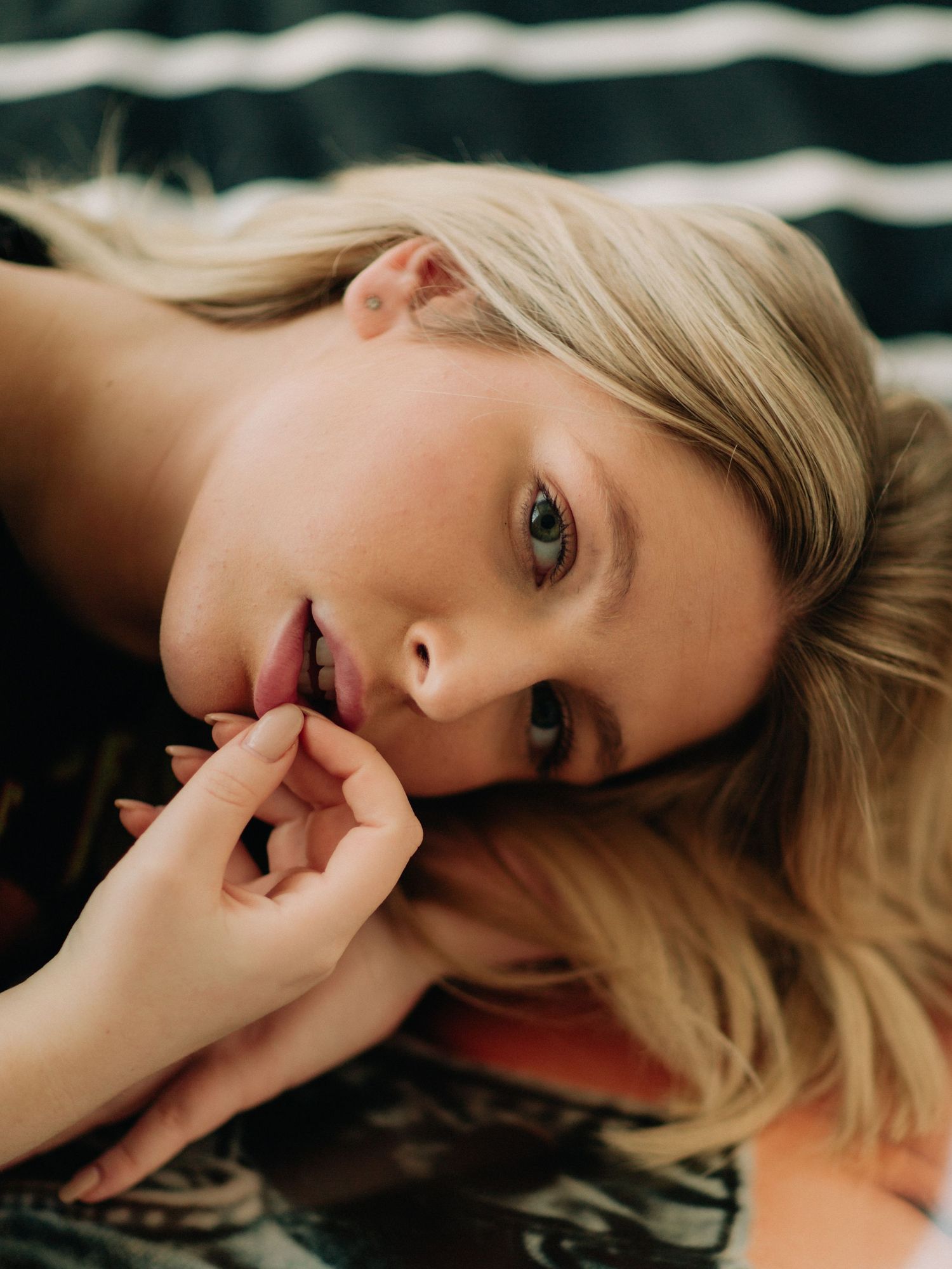 Young blonde woman lying down, gazing at camera with hand near lips, natural makeup, candid portrait photo.