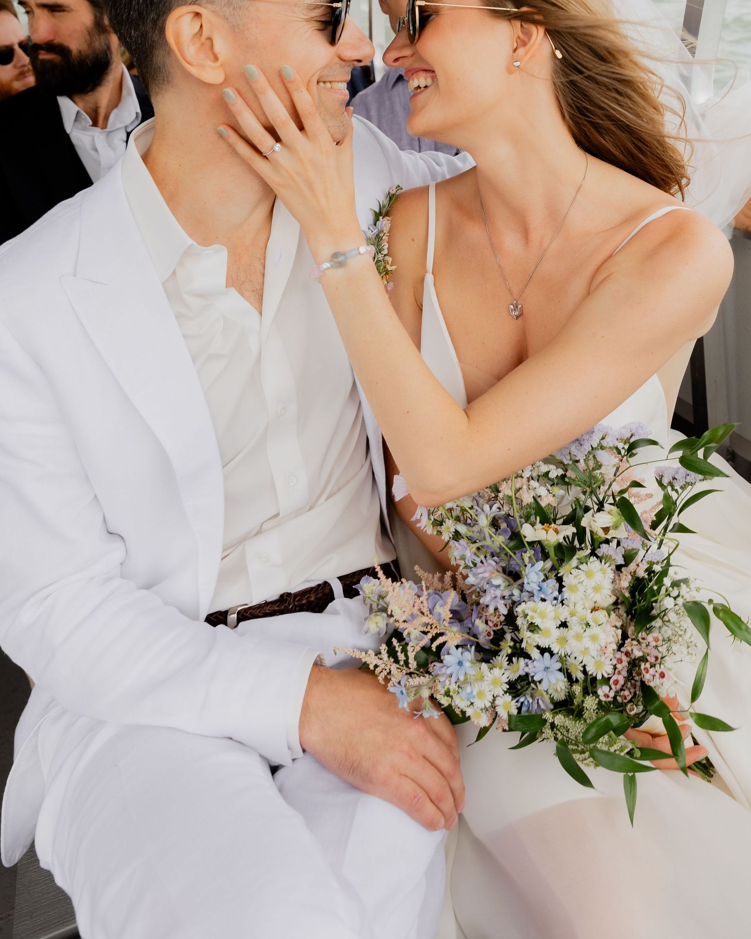 Romantic wedding couple embraces while holding a delicate bouquet of white and purple flowers against a light background.