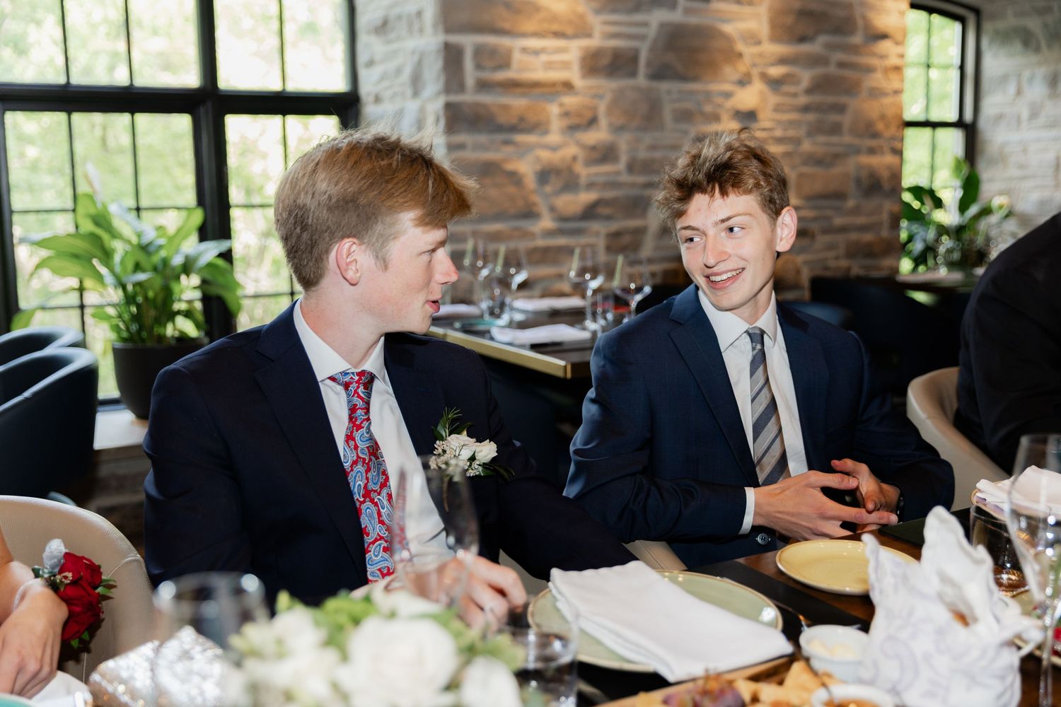 Two young men in suits and ties smiling at a wedding reception dinner table with floral arrangements.
