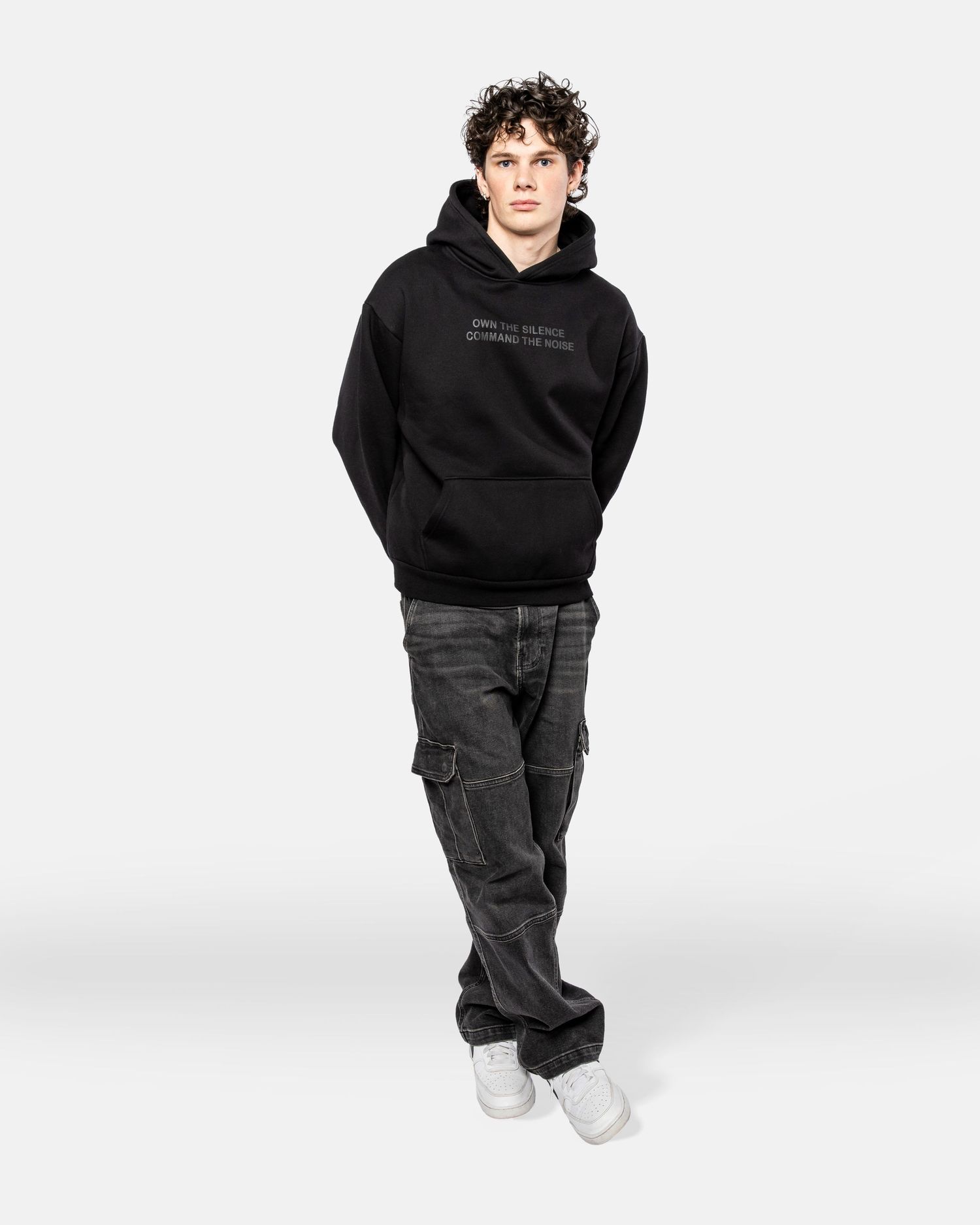 Person wearing a black hoodie and dark cargo pants stands against a white background with white sneakers.