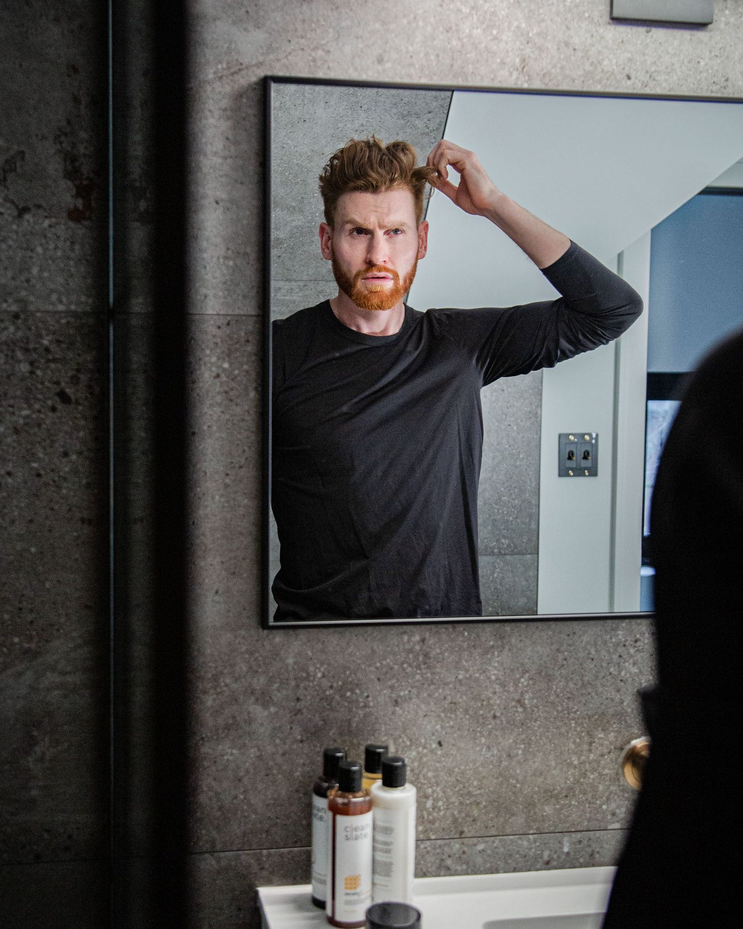 Person in dark shirt looking in mirror while applying grooming products.