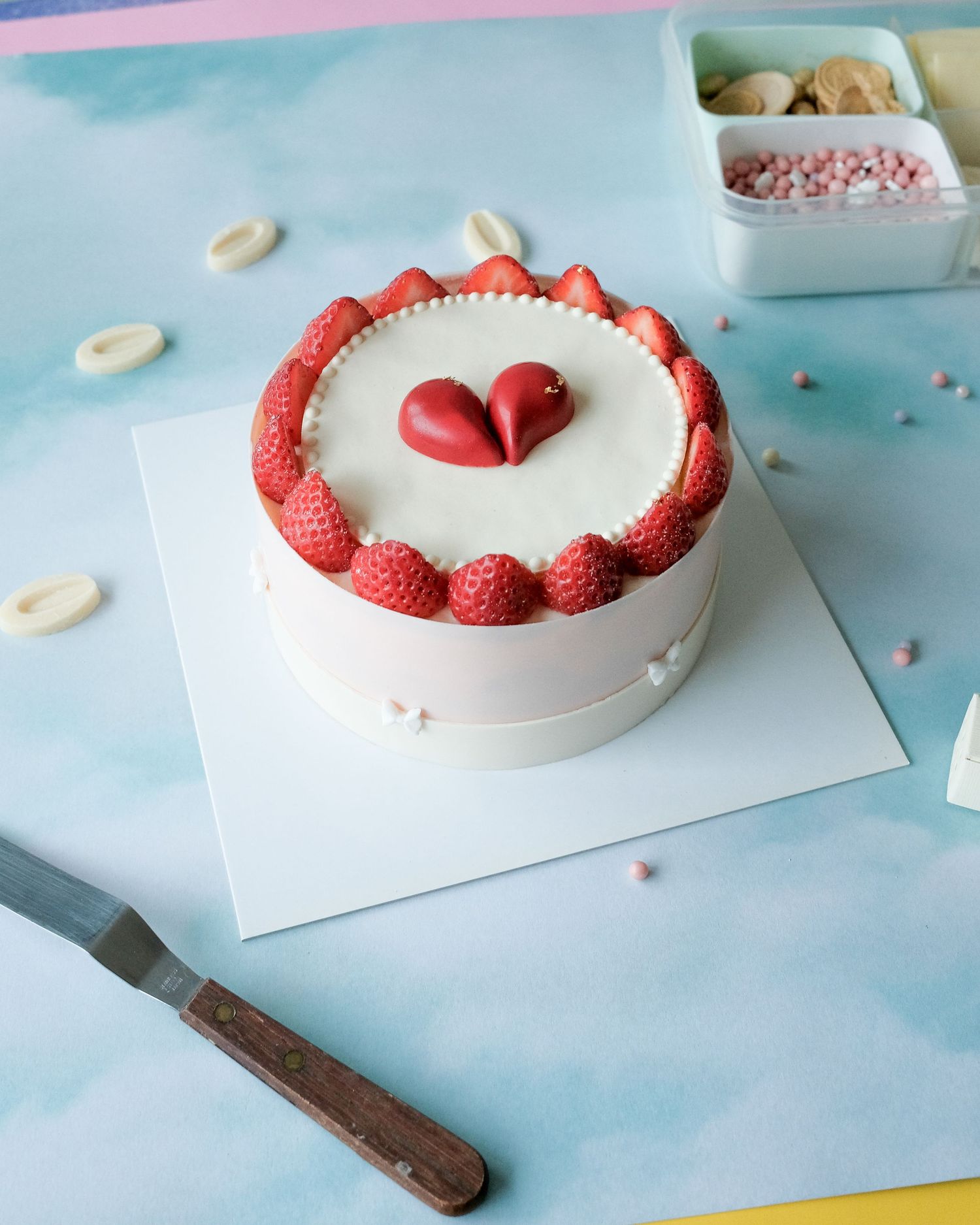 Round white cake decorated with fresh raspberries and red heart design on a light blue surface.