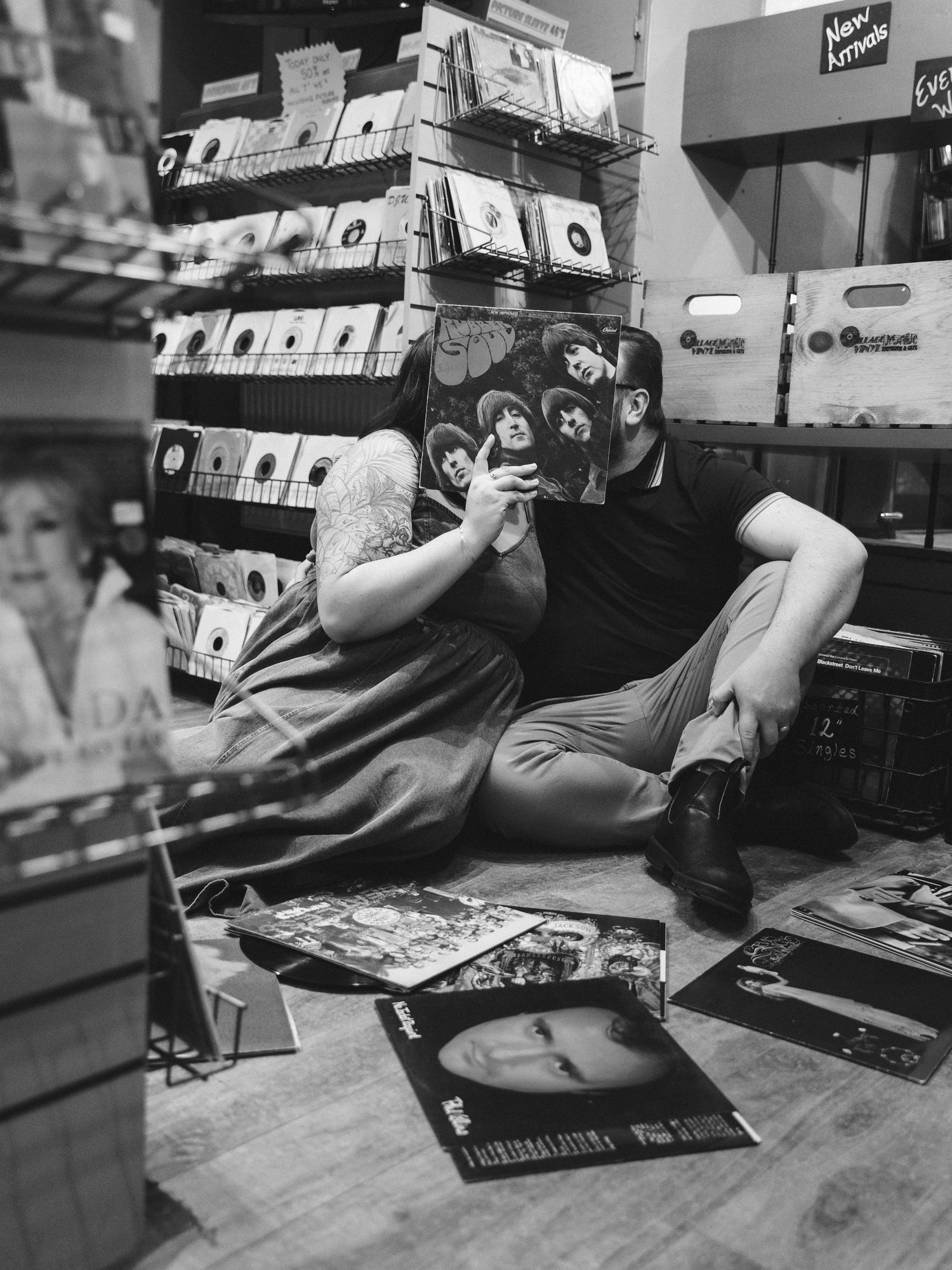 Black and white photo of a person sitting on a record store floor surrounded by vinyl albums.