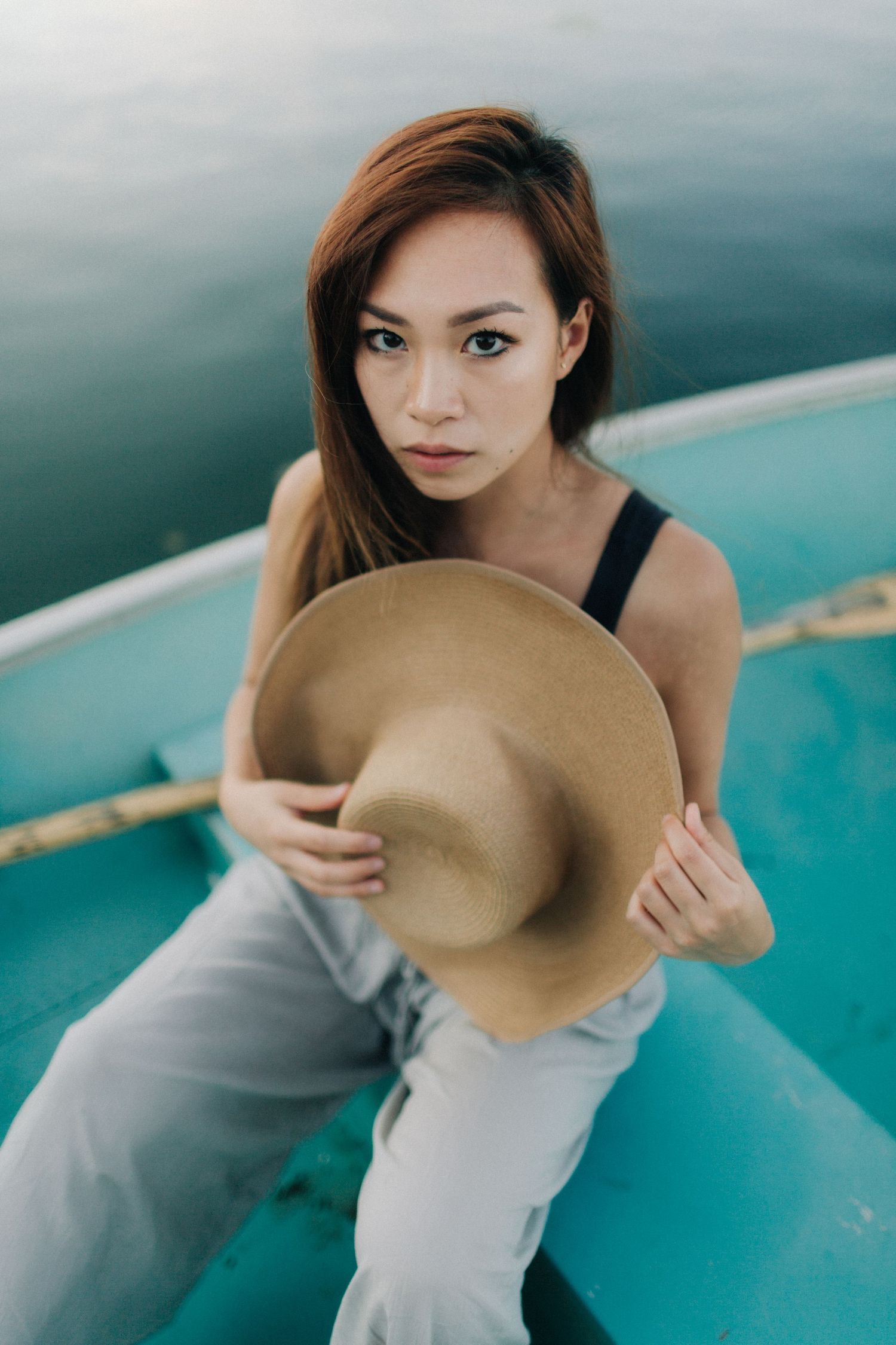 Young Asian woman holding a wide-brim tan hat while sitting in a turquoise boat near calm water.