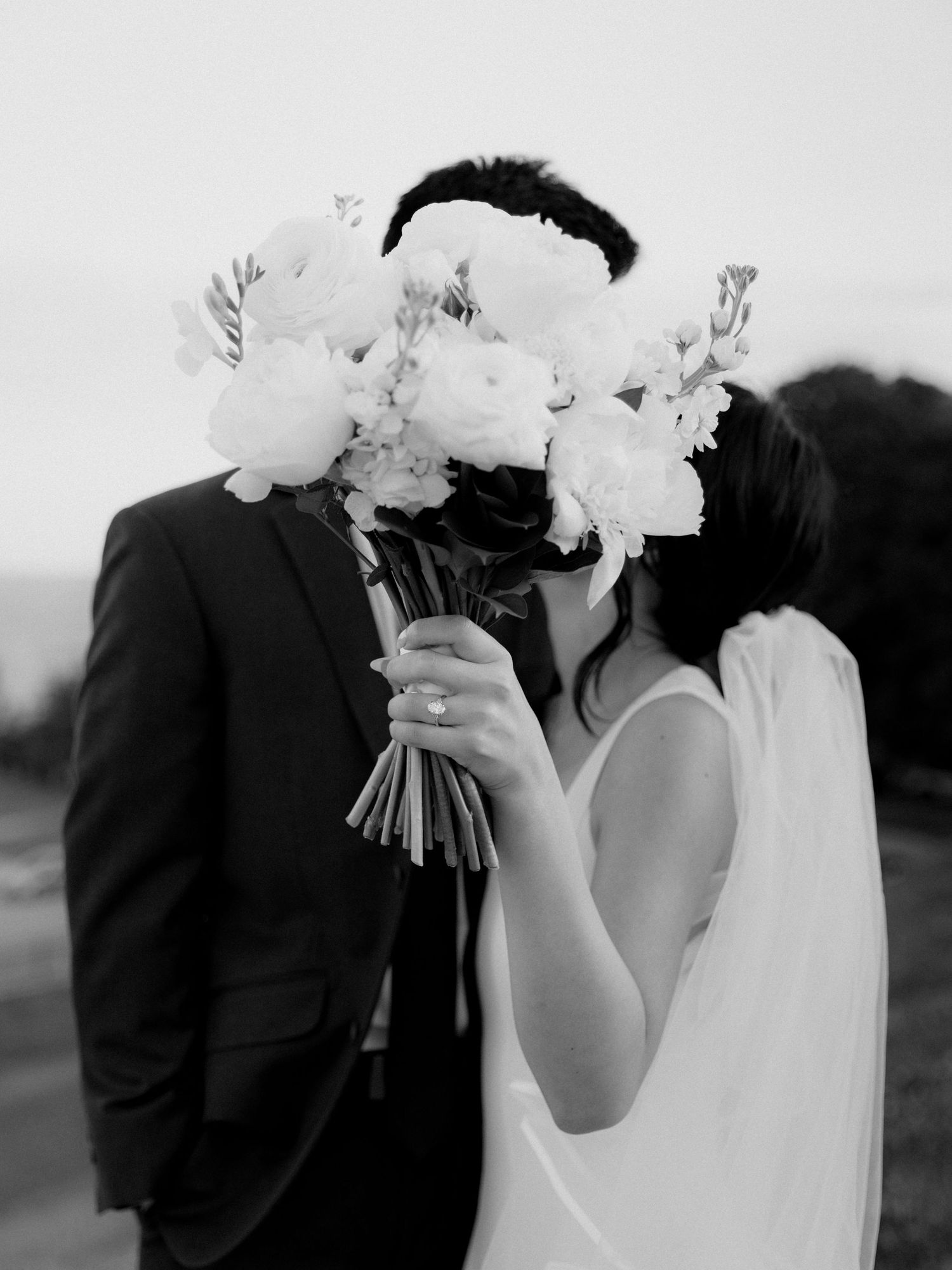 Black and white photo of a wedding couple kissing behind a white floral bouquet outdoors.