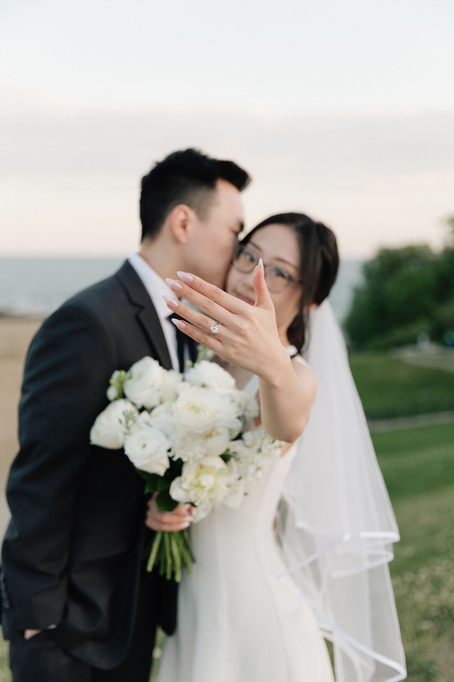Bride shows off wedding ring while groom kisses her cheek outdoors, holding white peony bouquet near waterfront.