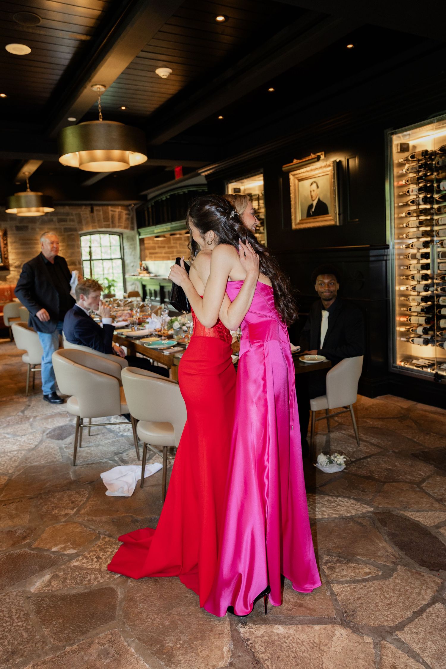 Two women in elegant red and pink gowns stand in a upscale restaurant dining room with wine display.