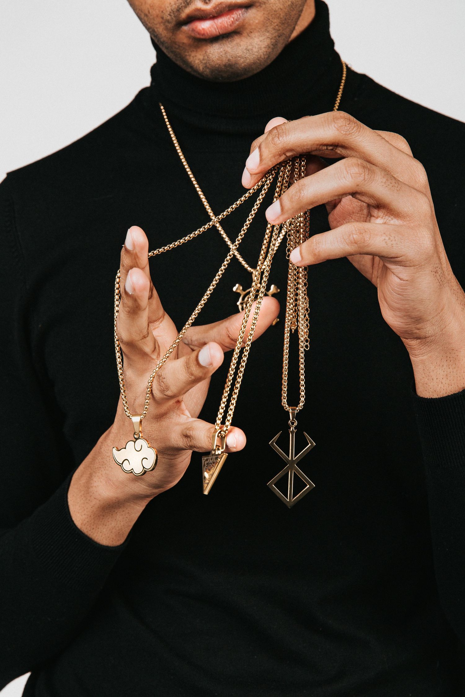 Hands displaying gold chain necklaces with pendants against black turtleneck.