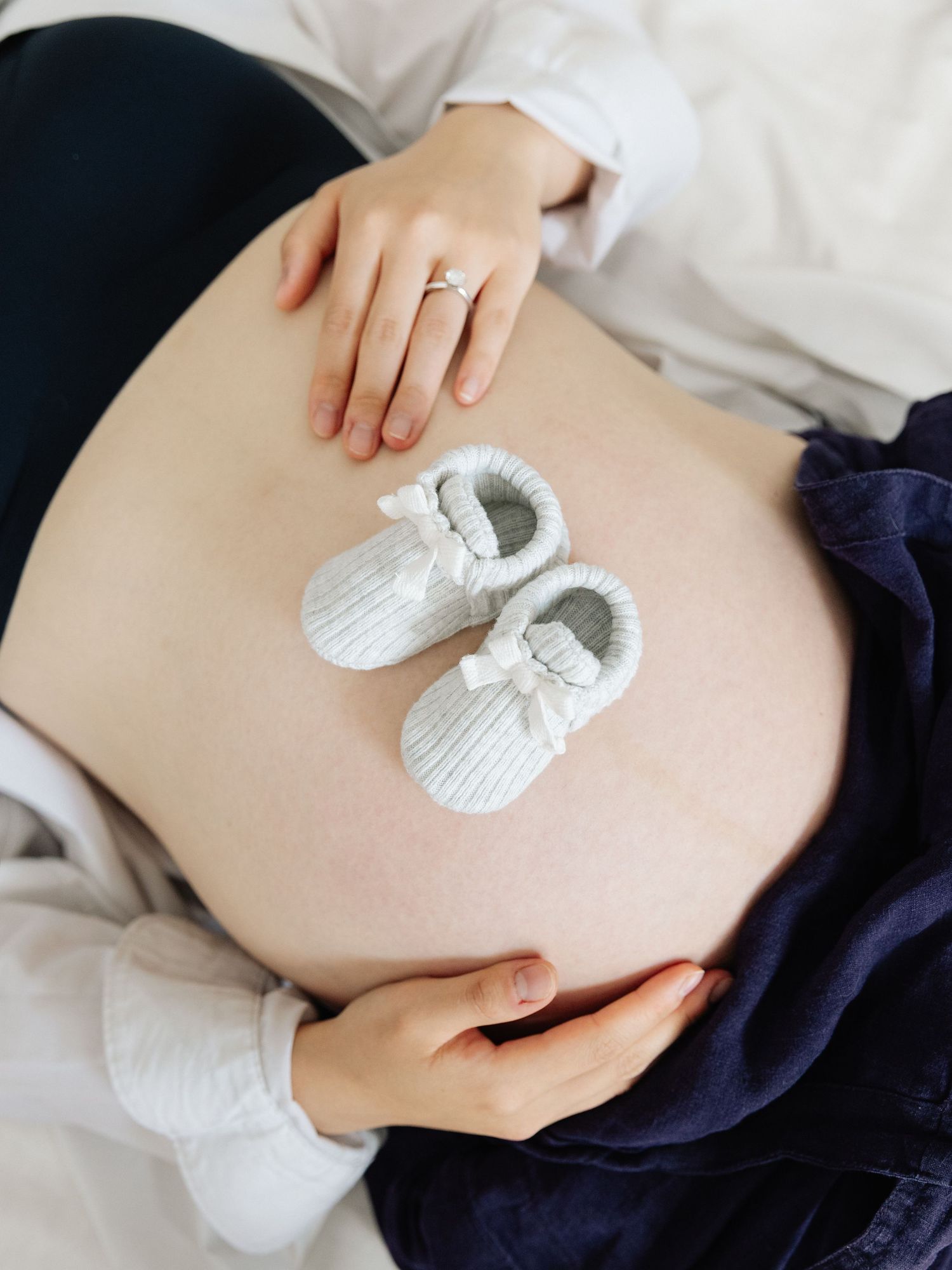 Pregnant woman resting with tiny white baby shoes placed on her bare belly, wearing a ring on her finger.