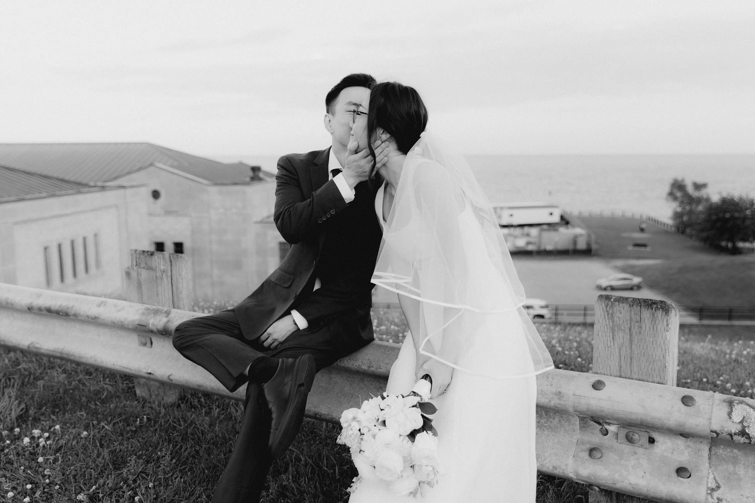 Black and white wedding photo of bride and groom sharing a kiss on a guardrail with ocean views.