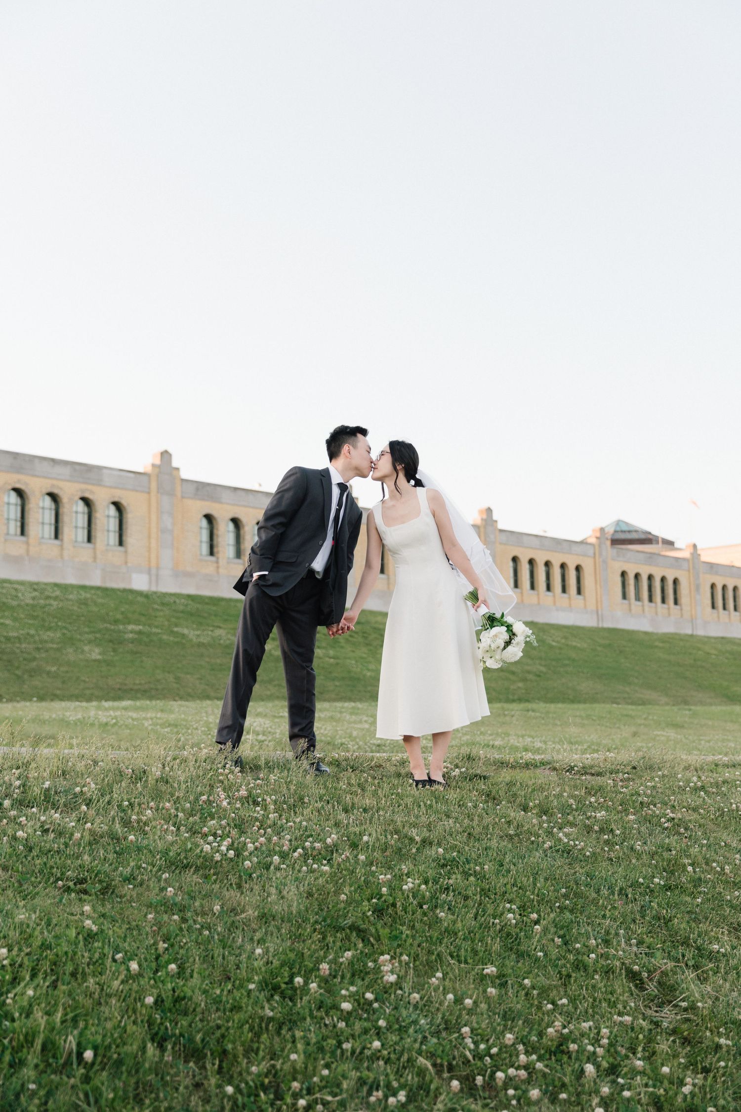 A couple shares a kiss on a green lawn, the groom in a black suit and bride in a white dress holding a bouquet.