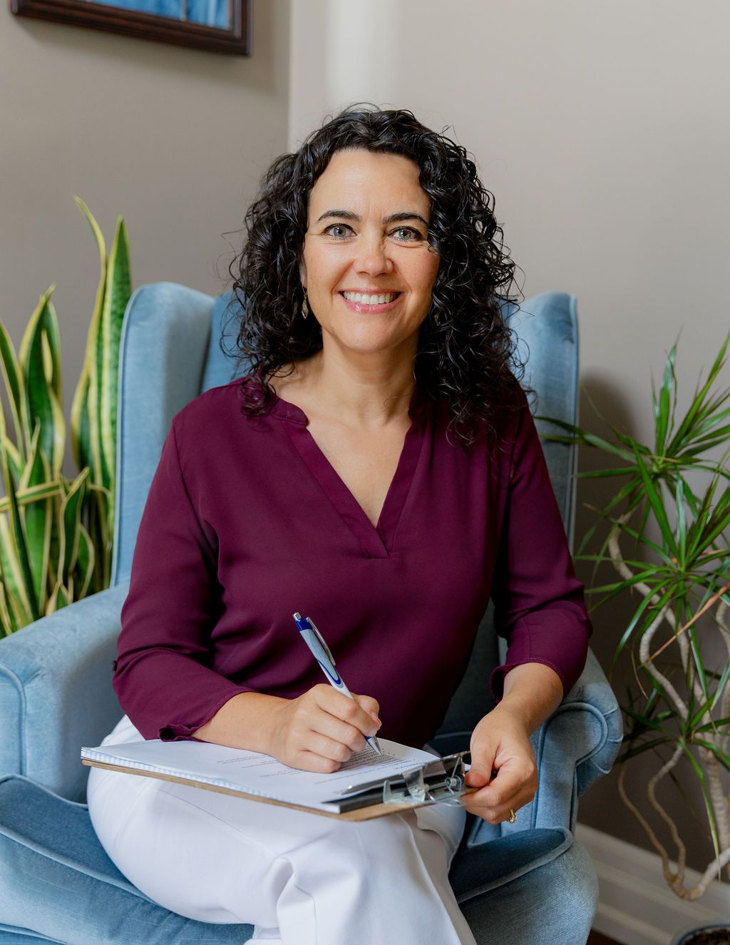 Smiling woman with curly hair sits in blue chair, writing on clipboard, wearing burgundy top, with plants in background.