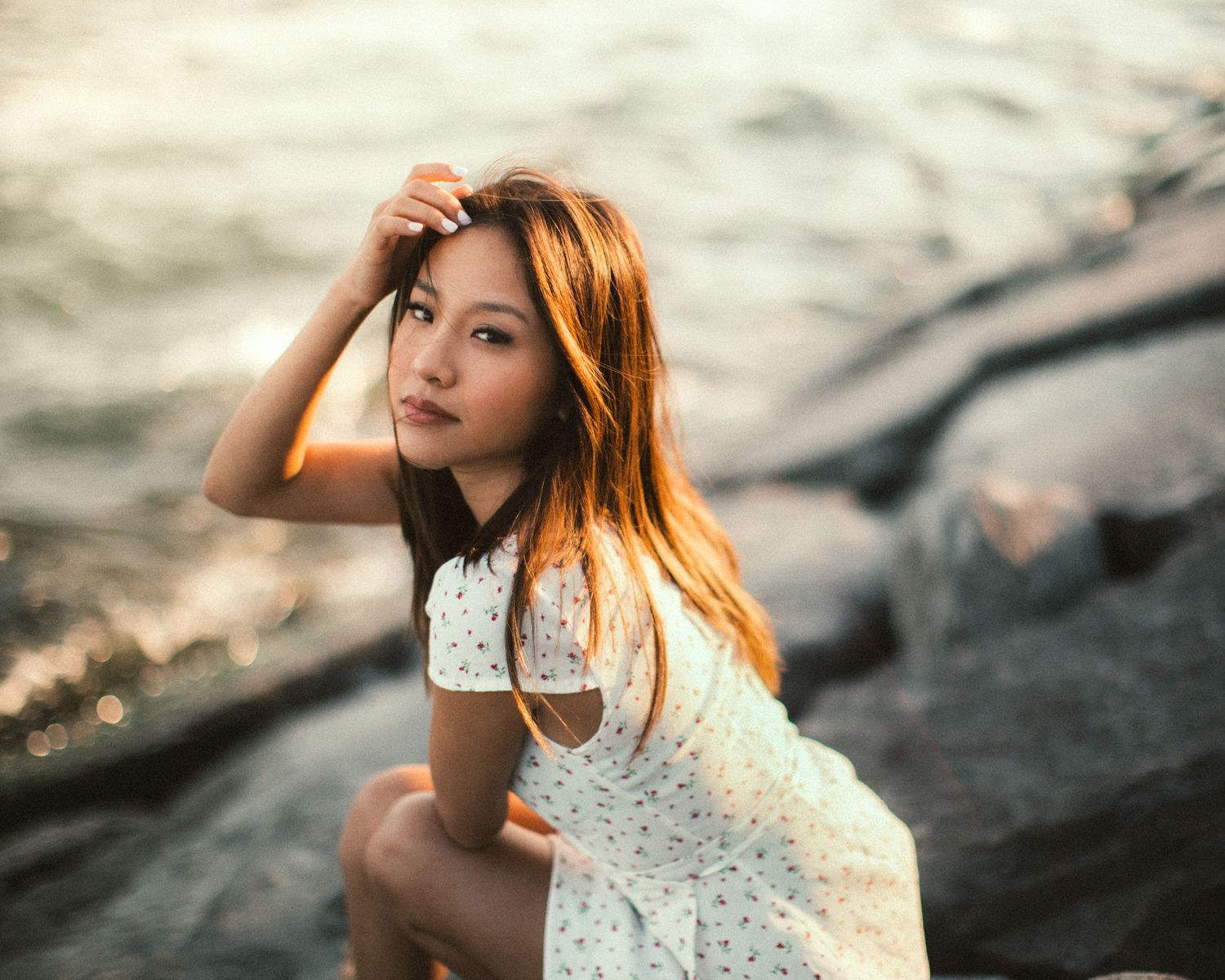 Artistic portrait of woman in white dress sitting on coastal rocks during golden hour.