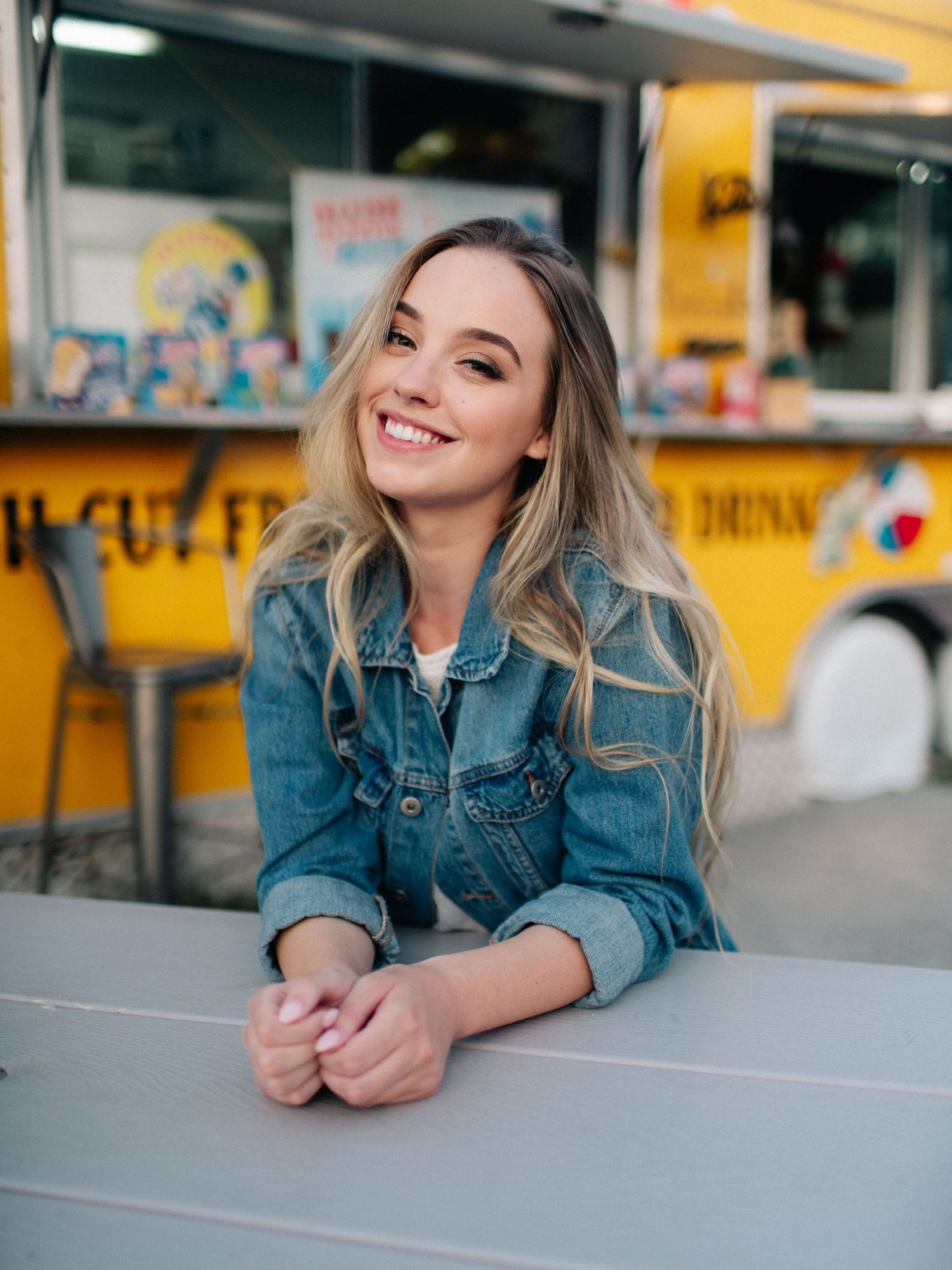 Smiling young woman with long blonde hair in denim jacket leaning on table near yellow food truck.