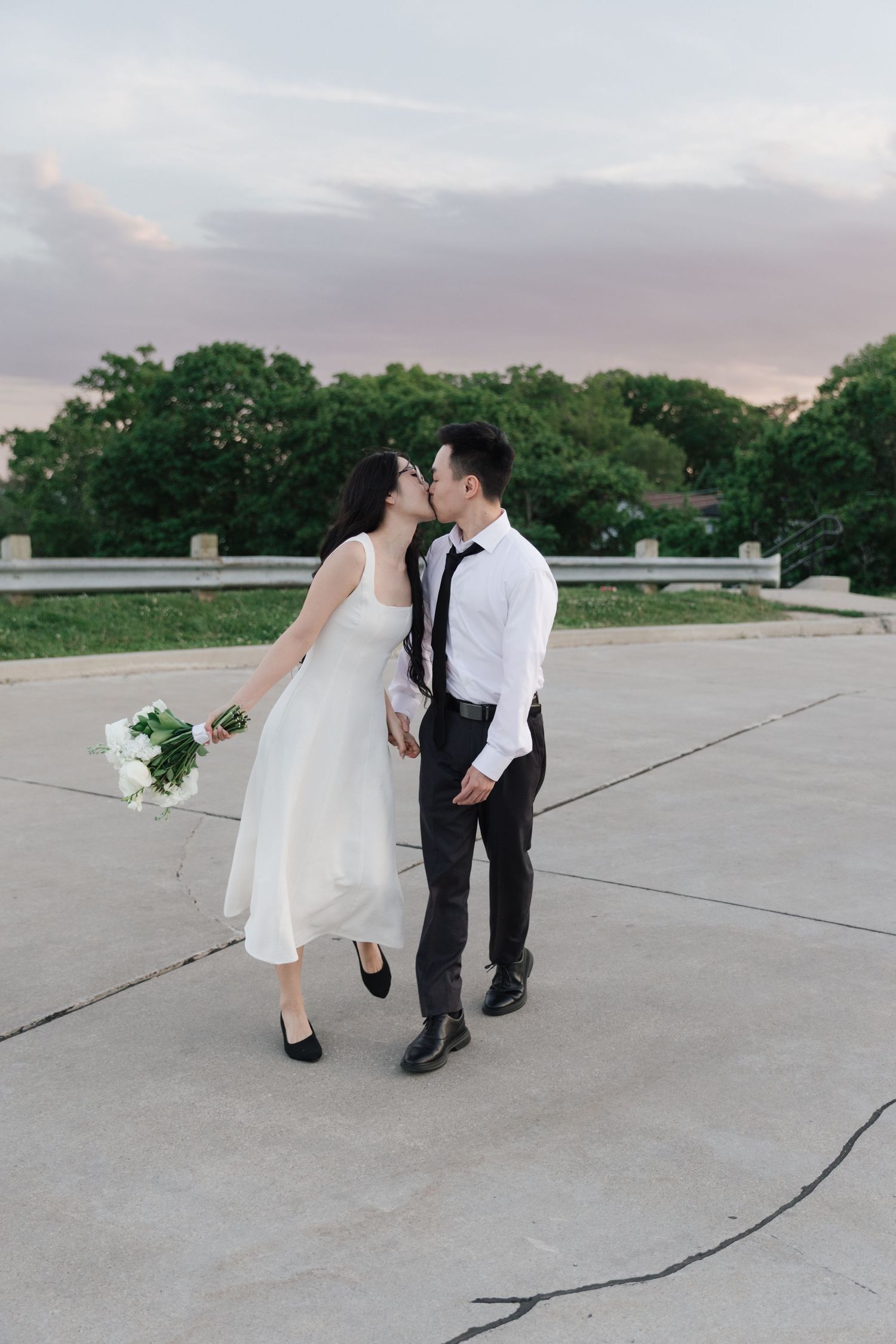 A couple in wedding attire shares a kiss while walking on a rooftop at sunset, bride holding white flowers.
