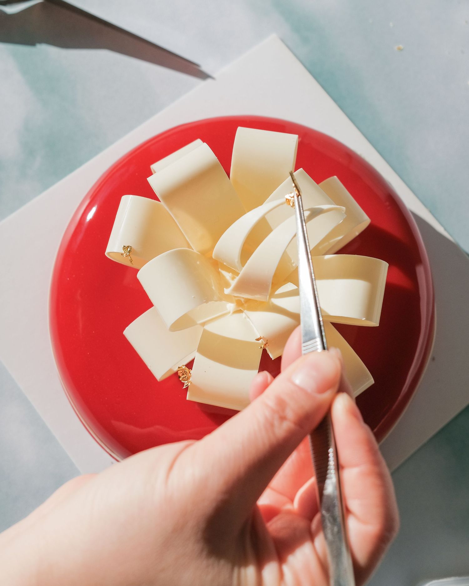 Hands carefully crafting white chocolate decorations on a red glazed cake surface.