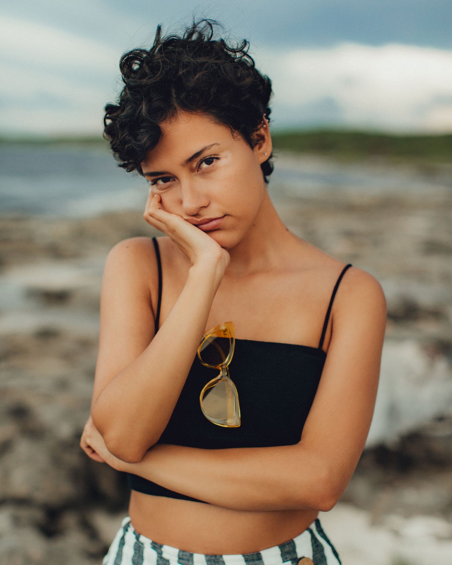 Fashion portrait of a model in black crop top and sunglasses against a coastal background.