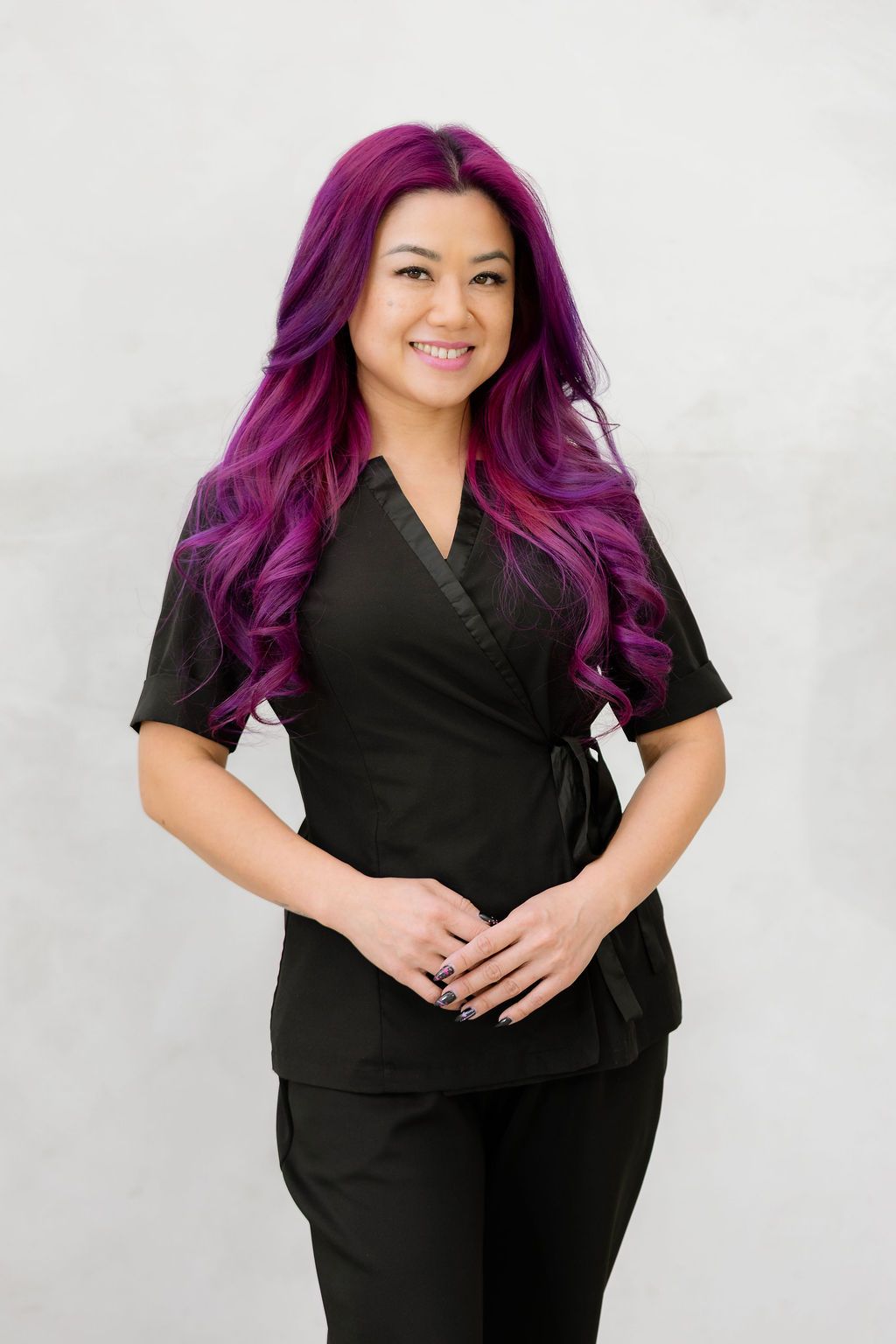 Smiling woman with long purple and pink hair wearing a black wrap top uniform against a gray background.