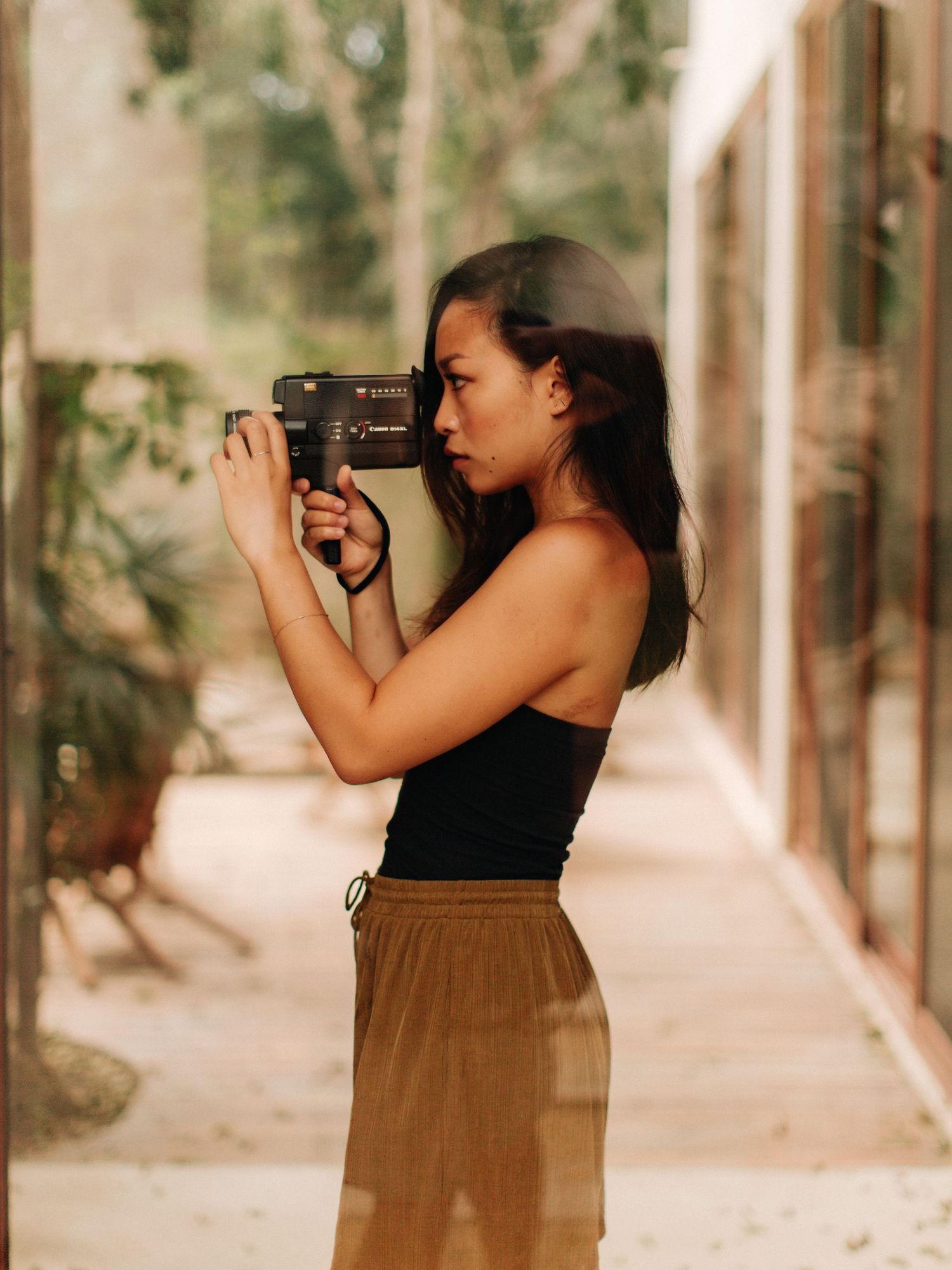 A young woman in a black tank top and olive skirt holds a vintage camera up to her eye outdoors.