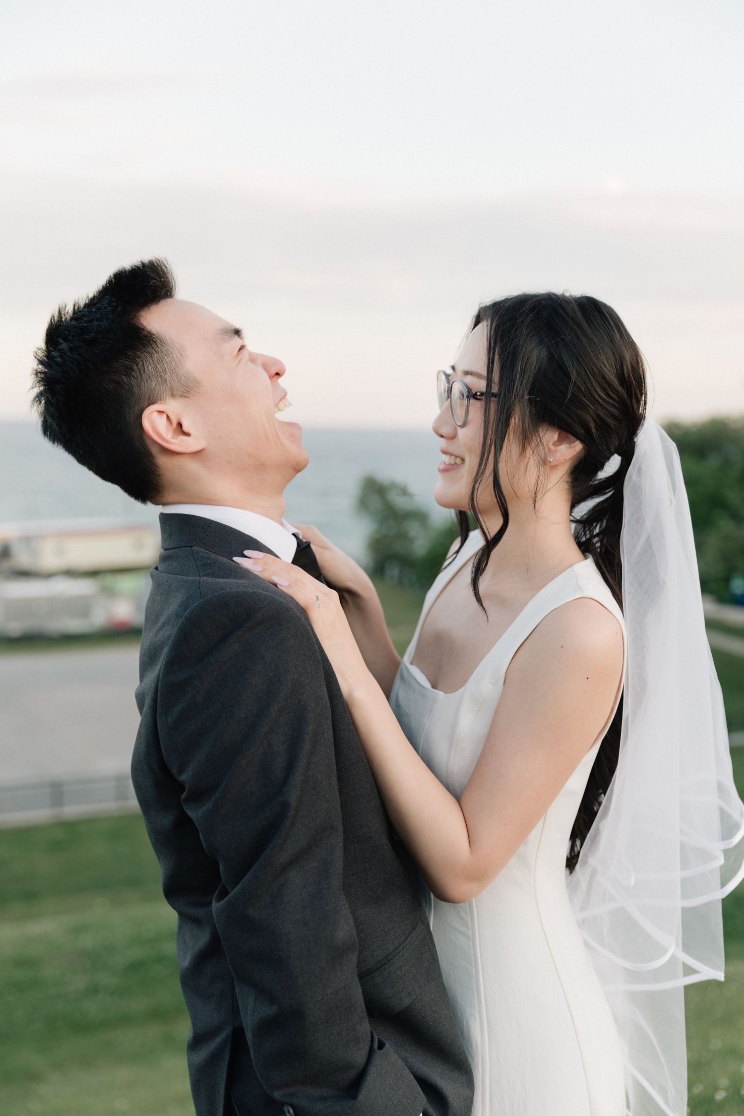 A smiling bride in a white gown and veil adjusts her groom's tie outdoors near the ocean at dusk.