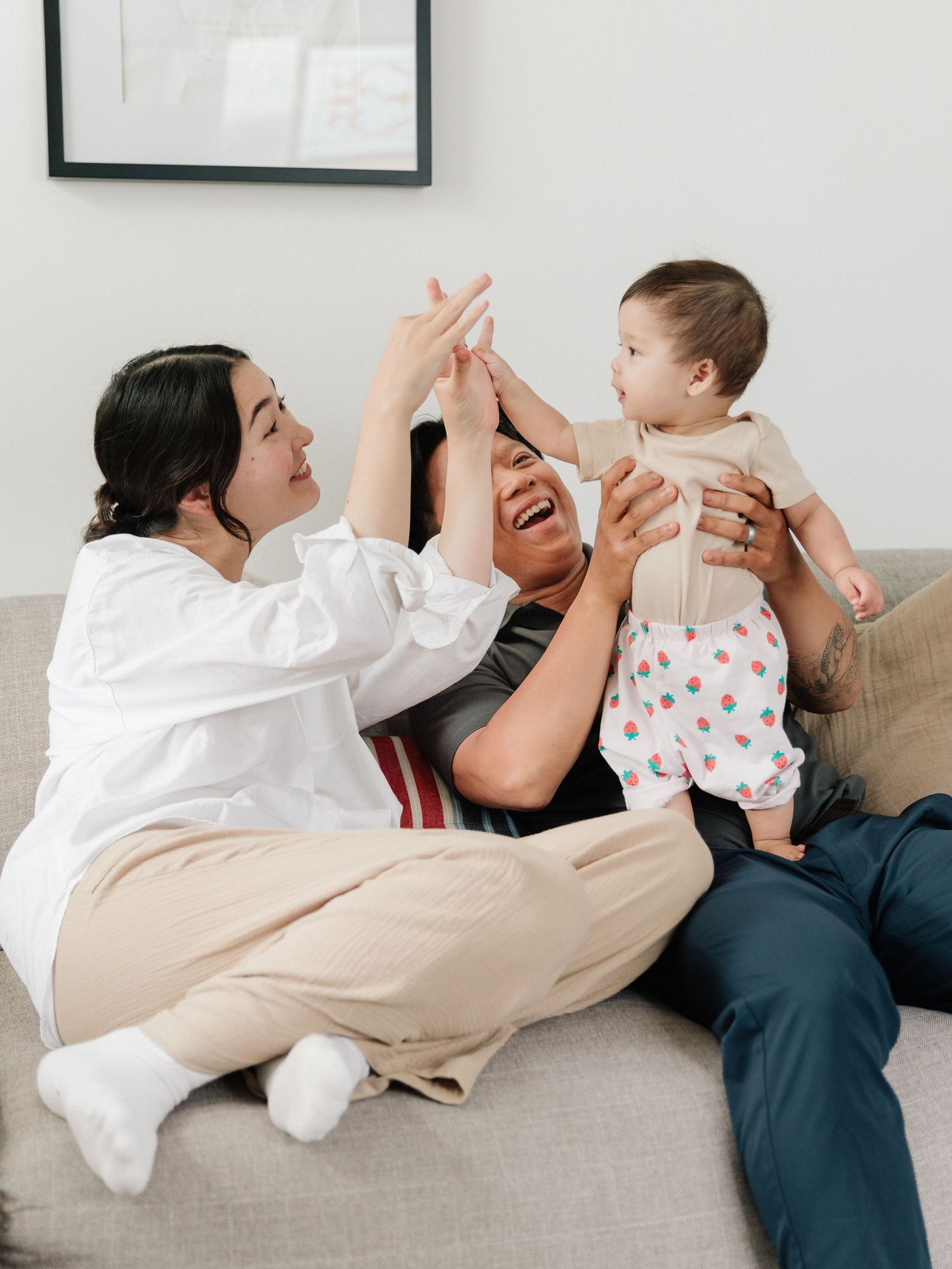 Two adults sitting on a couch joyfully playing with a smiling baby in a bright, cozy living room.