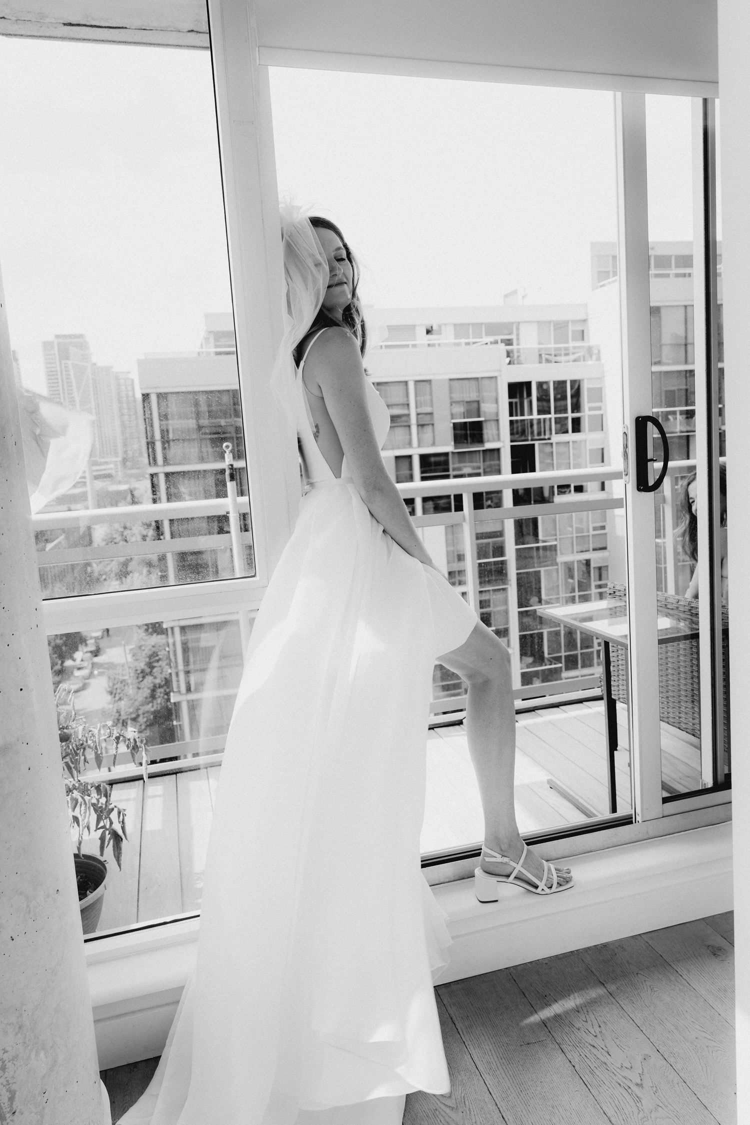 A black and white photo of a bride in a flowing white wedding dress standing near large windows in a bright room.