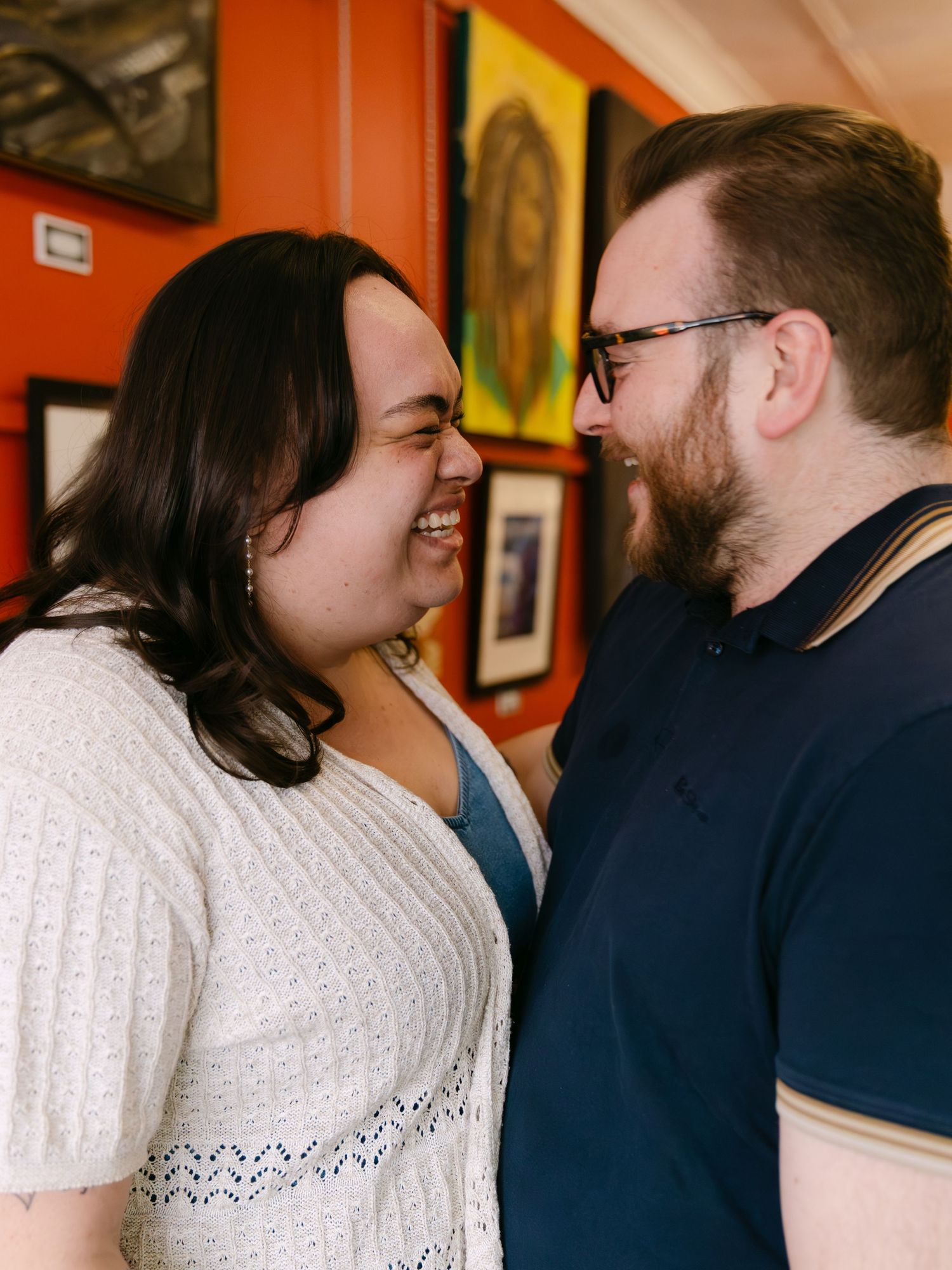 A smiling couple shares a joyful moment together in front of colorful artwork on an orange wall.