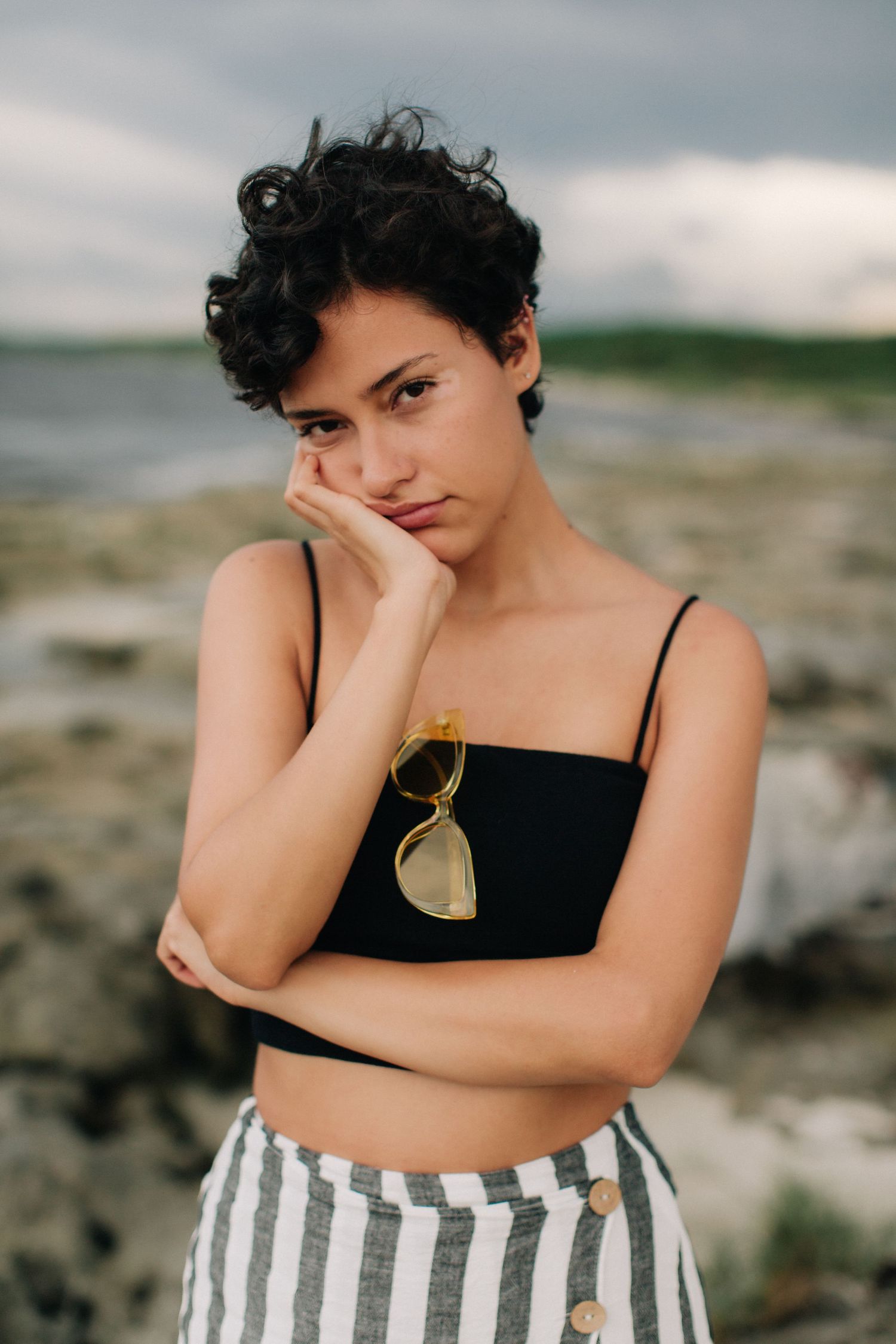 Young woman with short curly hair in black crop top and striped pants poses thoughtfully at rocky beach.