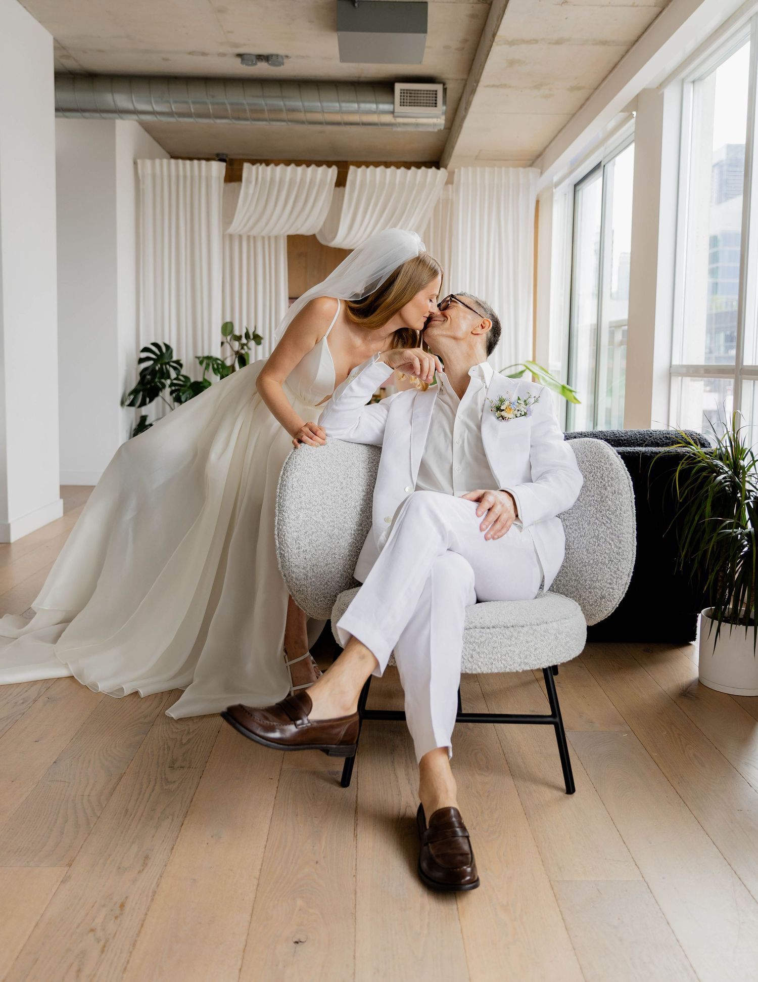 Bride in white gown kisses groom in white suit seated in modern chair by large windows on wedding day.