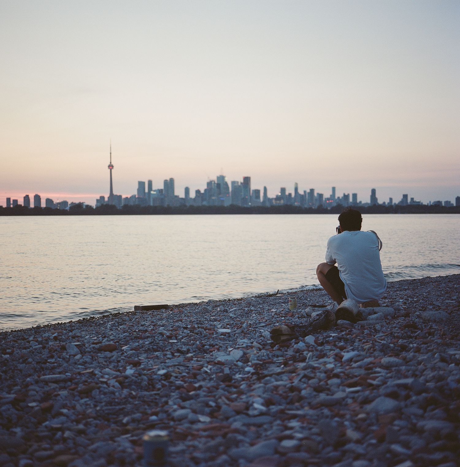 A solitary figure sits on a rocky shore overlooking Toronto's iconic skyline at sunset.