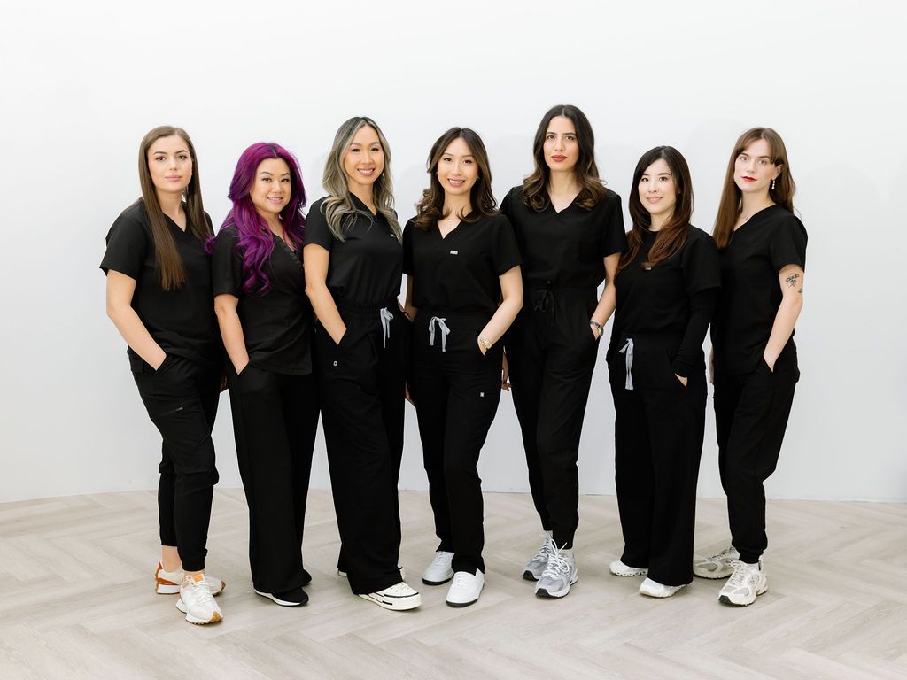 Seven women in matching black scrubs pose together in a bright, modern medical or spa clinic setting.