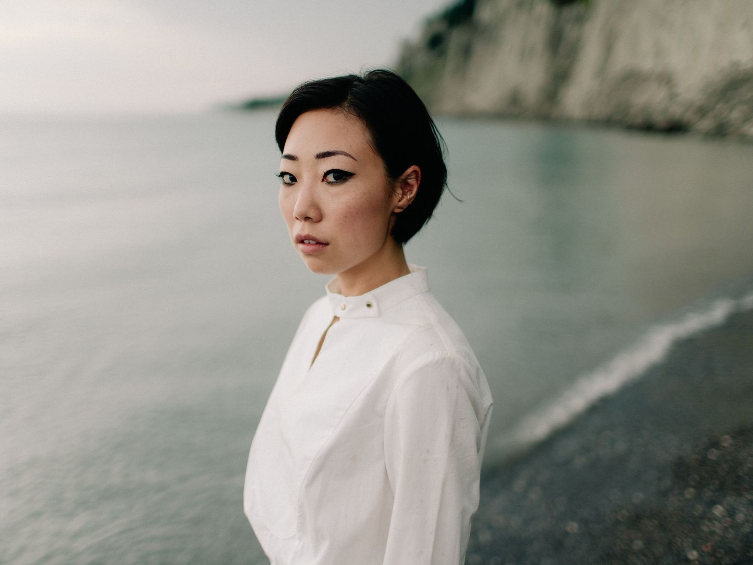 A young Asian woman in a white blouse stands on a rocky beach with calm ocean waters and cliffs behind her.