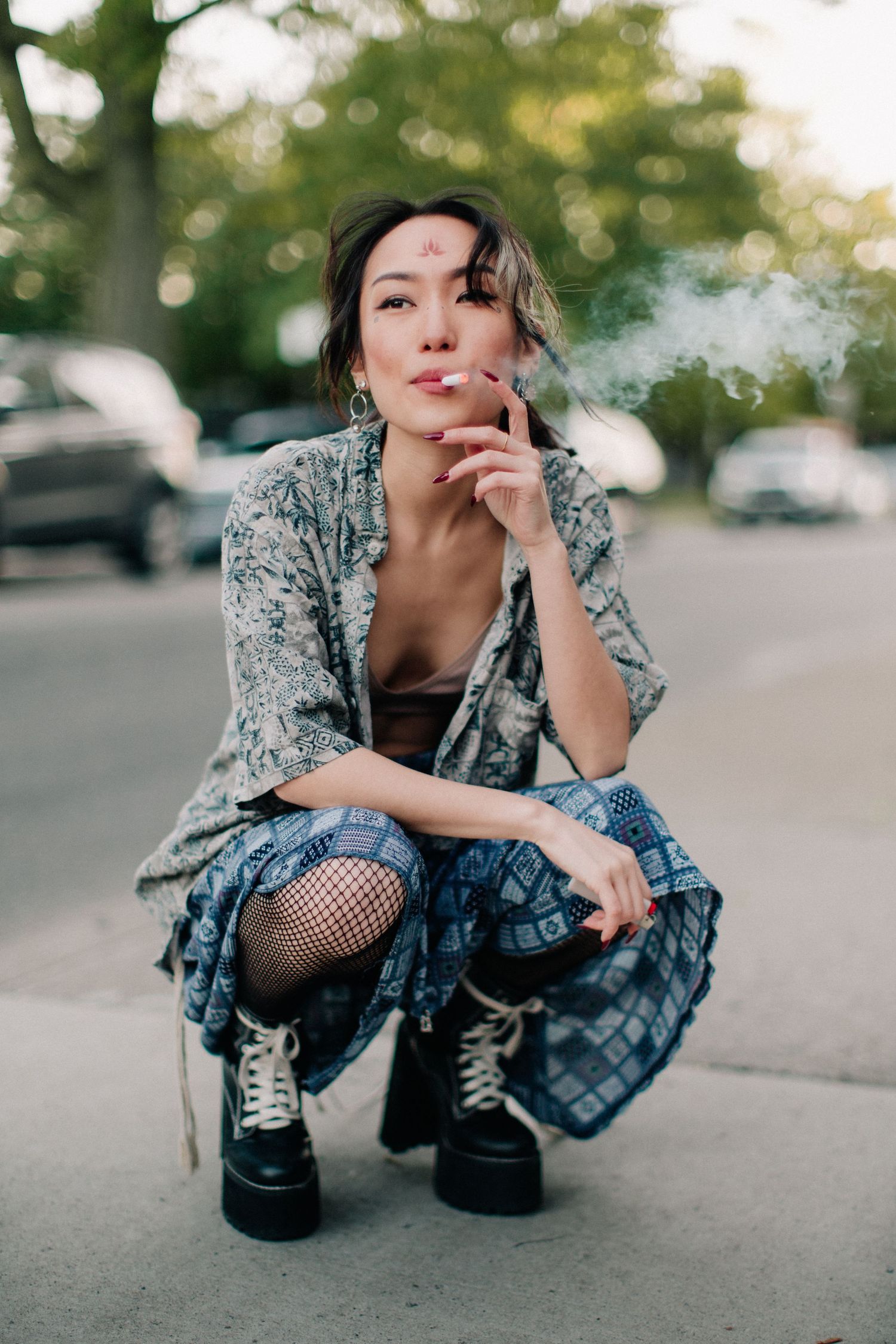 A woman in bohemian attire with fishnet tights and platform boots crouches on a sidewalk while smoking a cigarette.