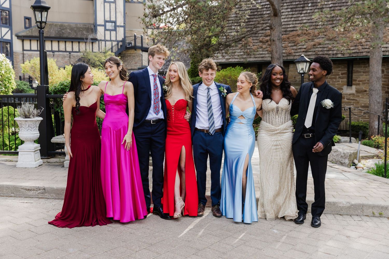 Group of teens in formal prom attire posing together outdoors, wearing elegant gowns and suits.