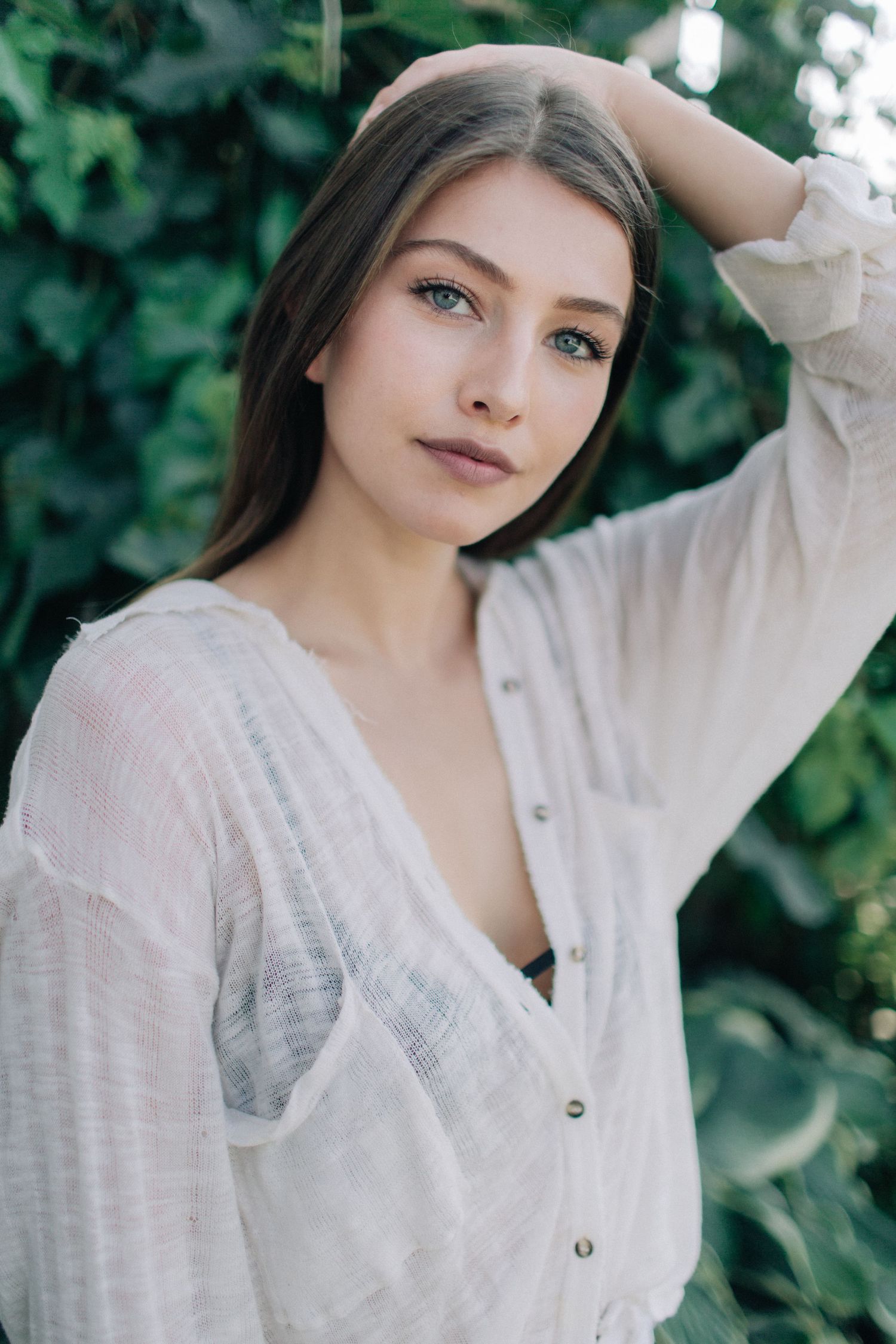 Young woman with long brown hair and blue eyes in a white knit cardigan posing outdoors near green foliage.