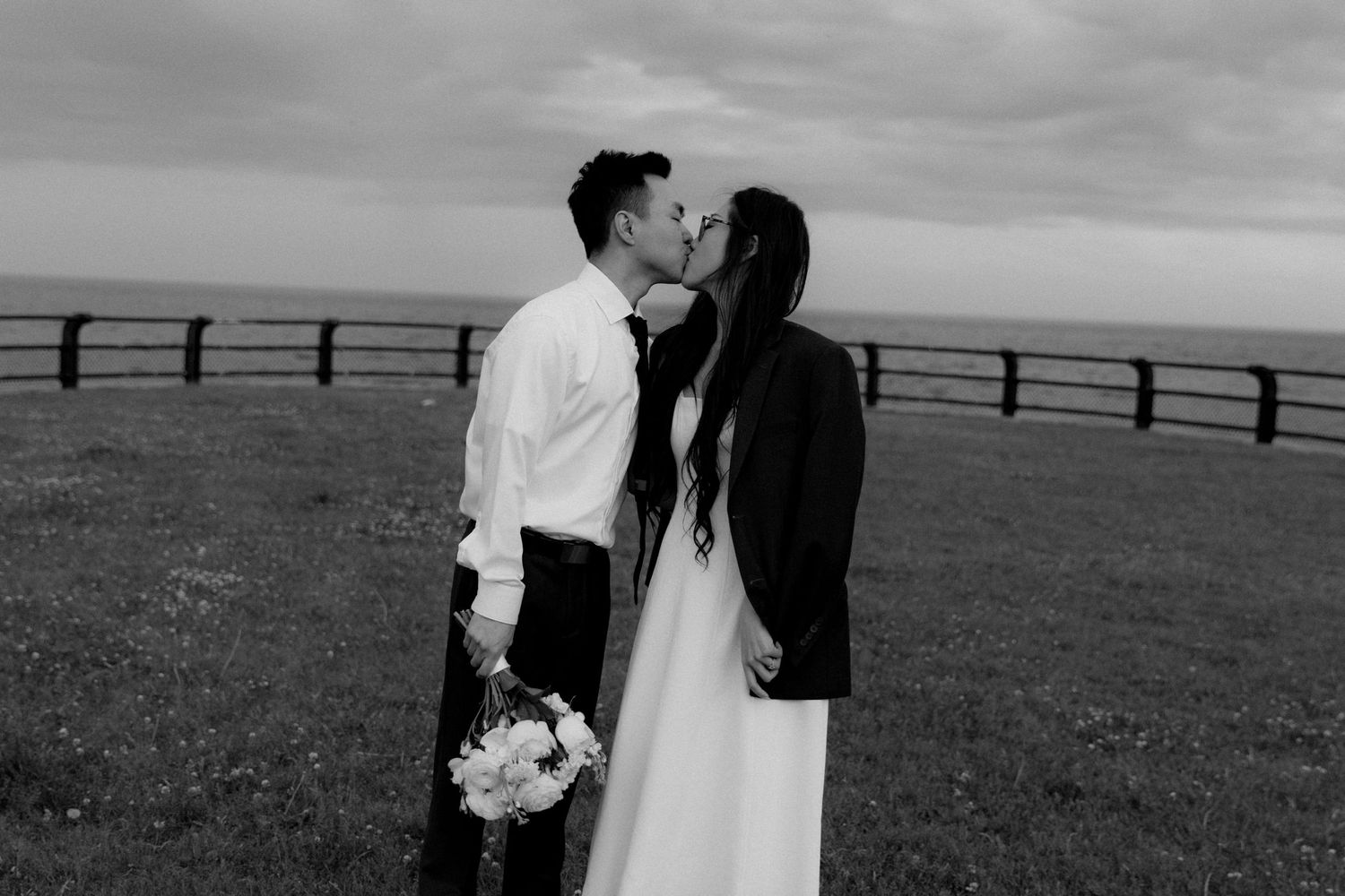 Black and white photo of a bride and groom sharing a kiss in a grassy field with a wooden fence and ocean backdrop.