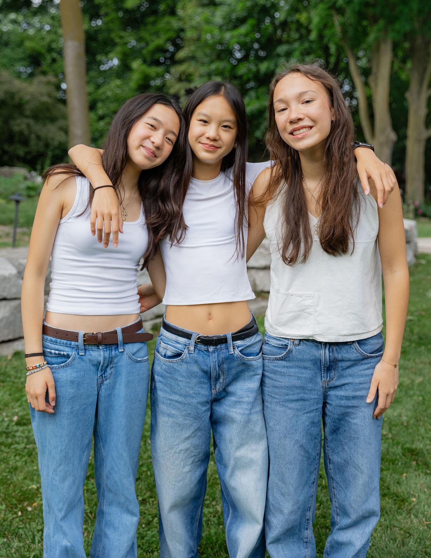 Friends in matching white tops and blue jeans pose casually outdoors in a park setting.