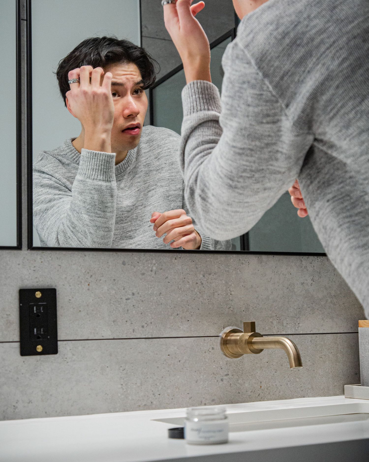 Person in grey sweater applying skincare product in front of bathroom mirror.