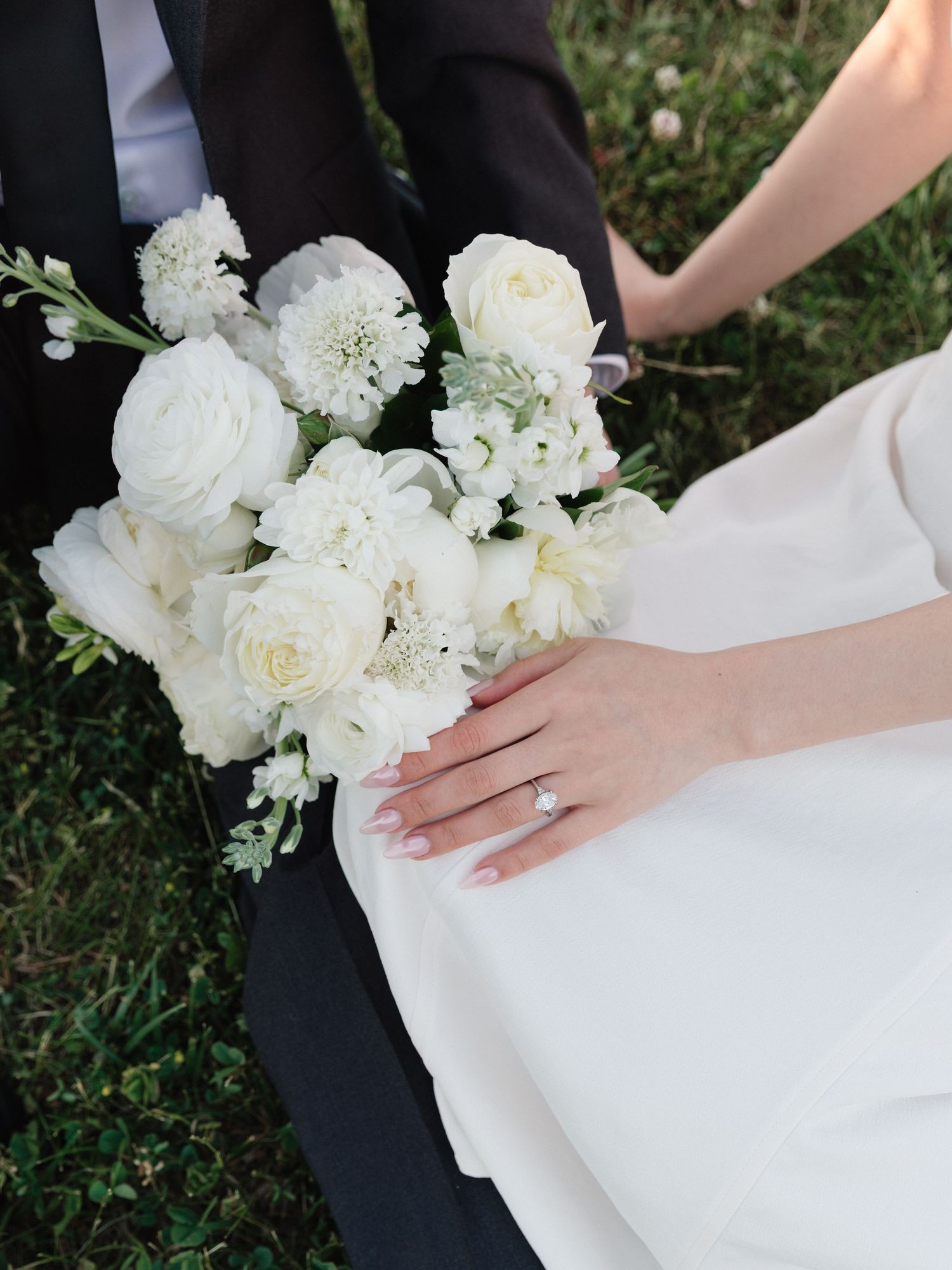 Bride and groom holding a white floral bouquet with roses, with bride showcasing diamond engagement ring.