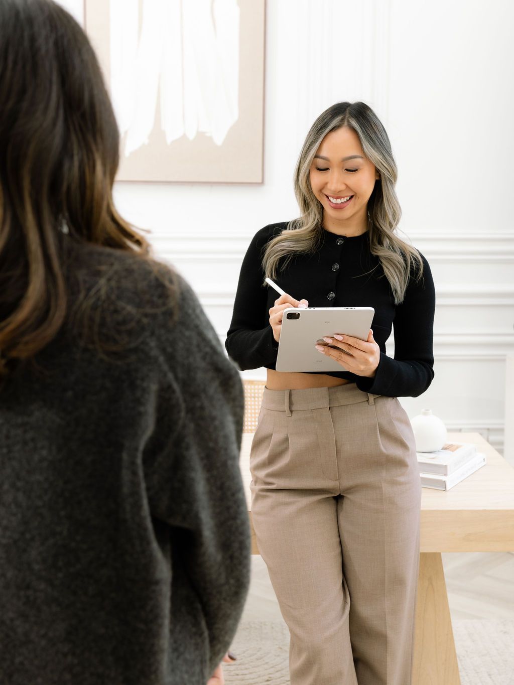 A smiling professional woman holds a tablet while consulting with a colleague in a modern office setting.
