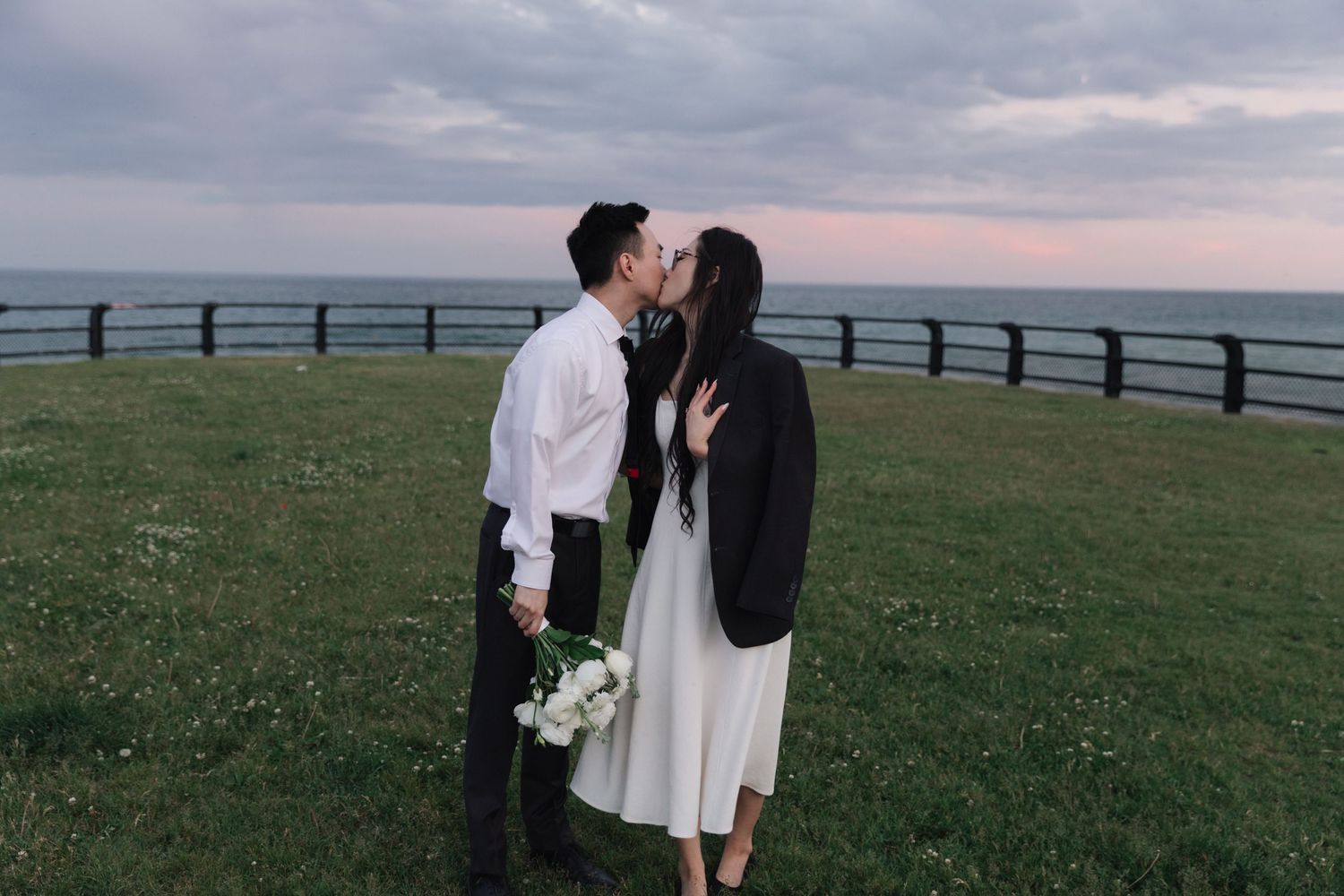 Couple sharing a romantic kiss on a grassy cliff overlooking the ocean at sunset, dressed in wedding attire.