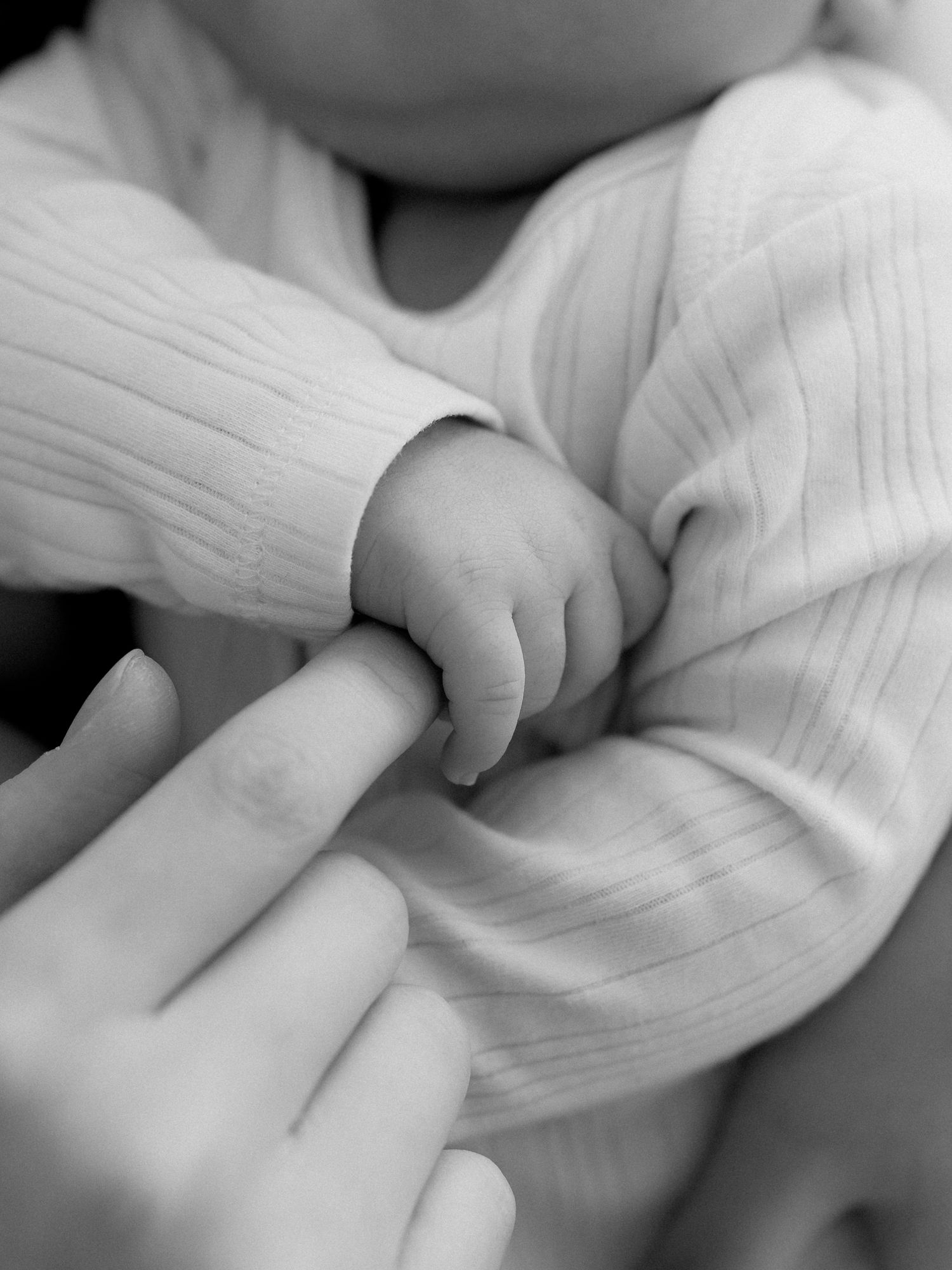 Black and white photo of a newborn baby grasping an adult's finger, wearing a soft ribbed onesie.