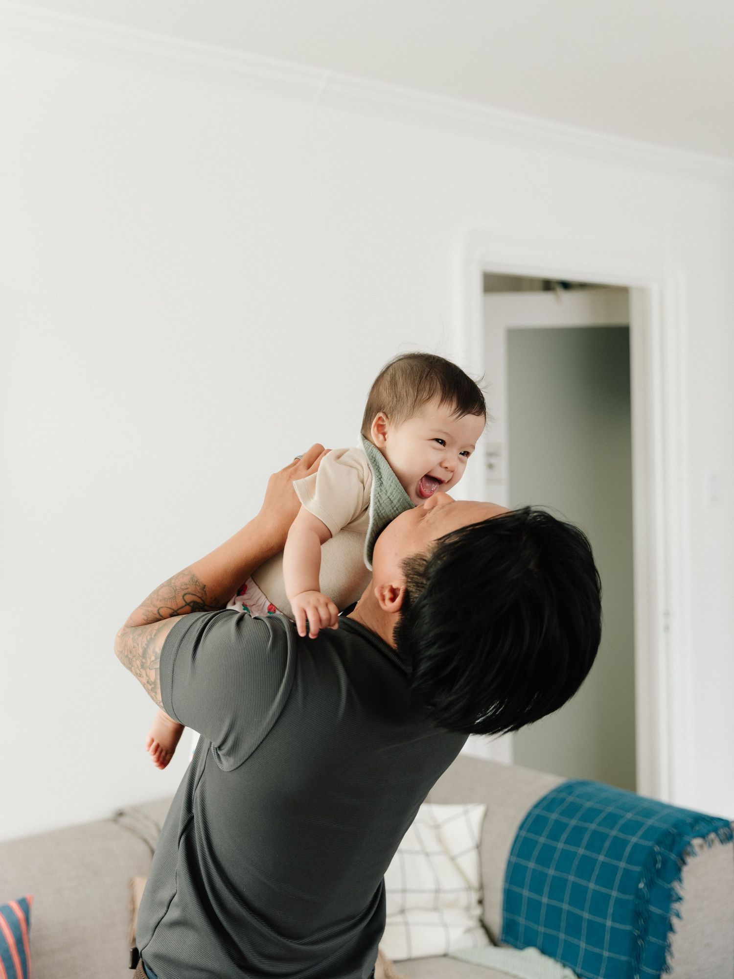 A father joyfully lifts his laughing baby above his head in a bright living room.