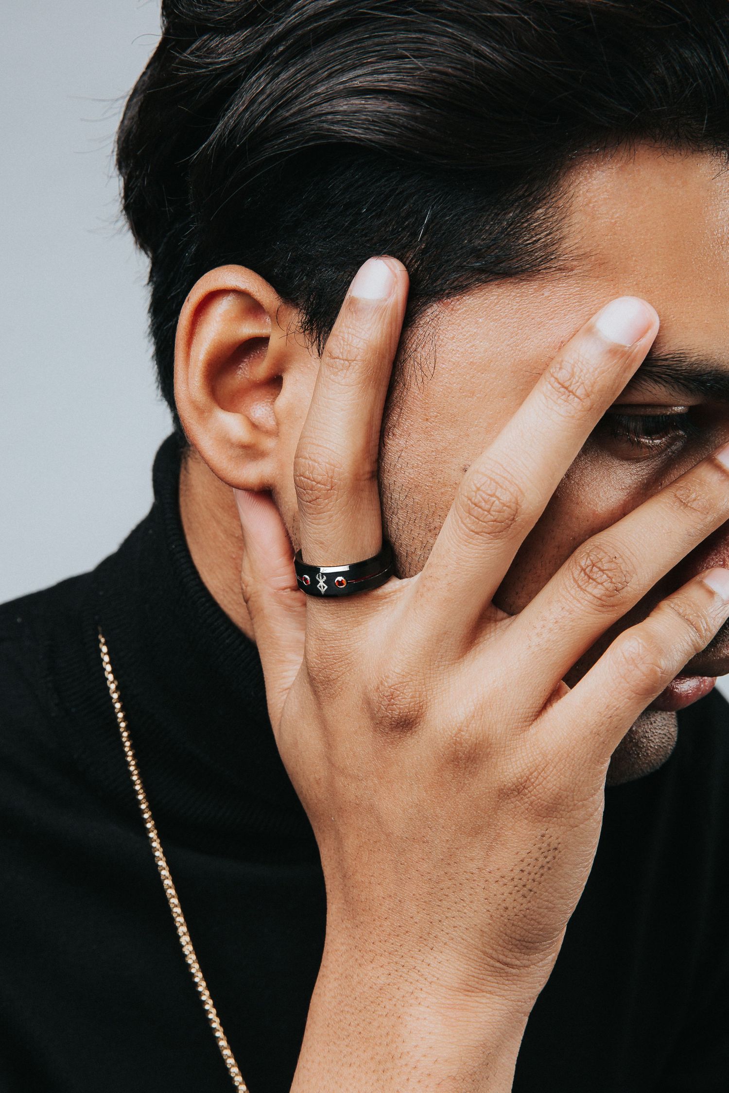 Artistic portrait showing hand covering face while wearing black ring and gold chain necklace.