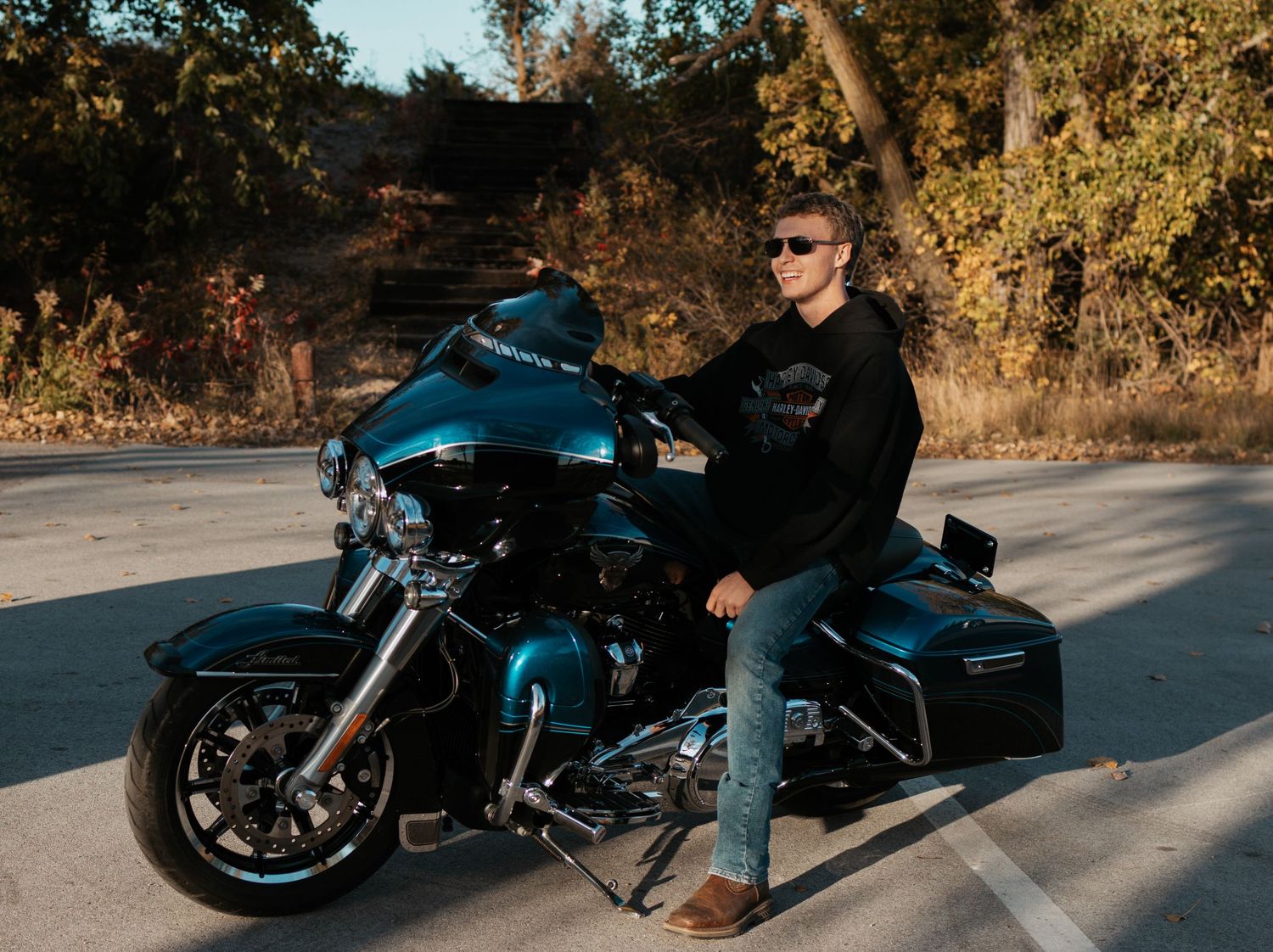 Young man in Harley-Davidson hoodie posing on teal motorcycle on open road with autumn trees.