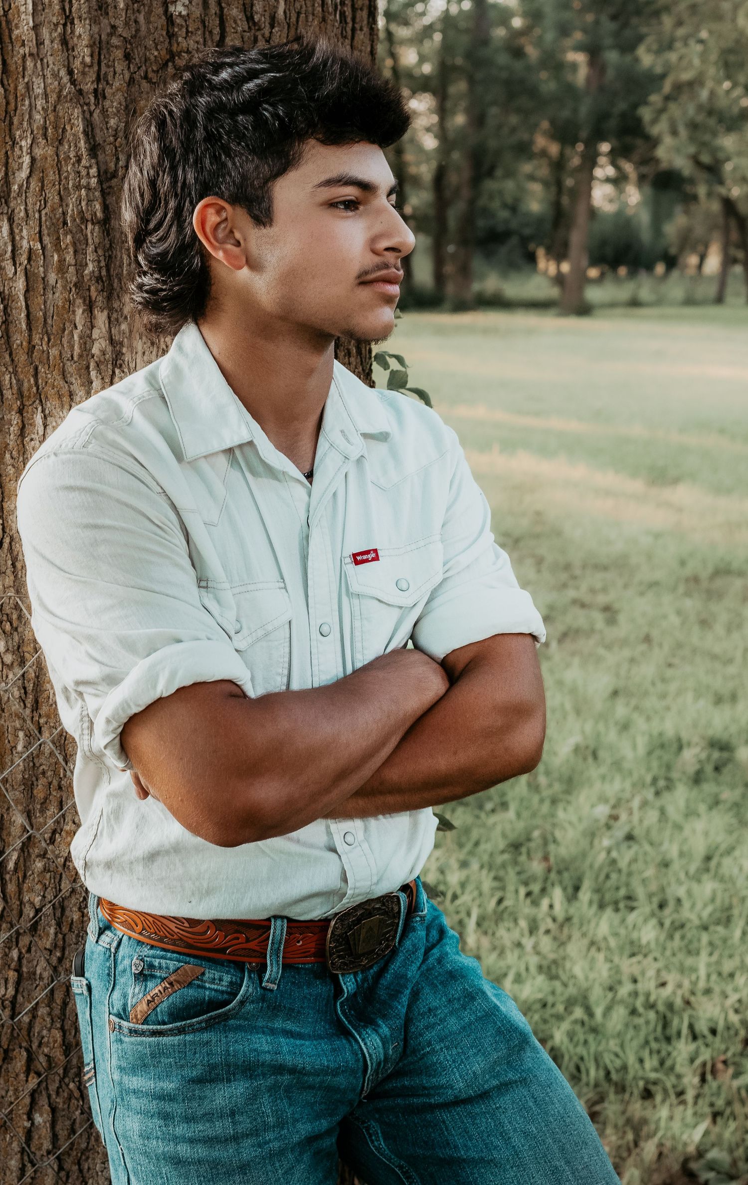 Young man in white Wrangler shirt and jeans leaning against tree with arms crossed in outdoor setting.