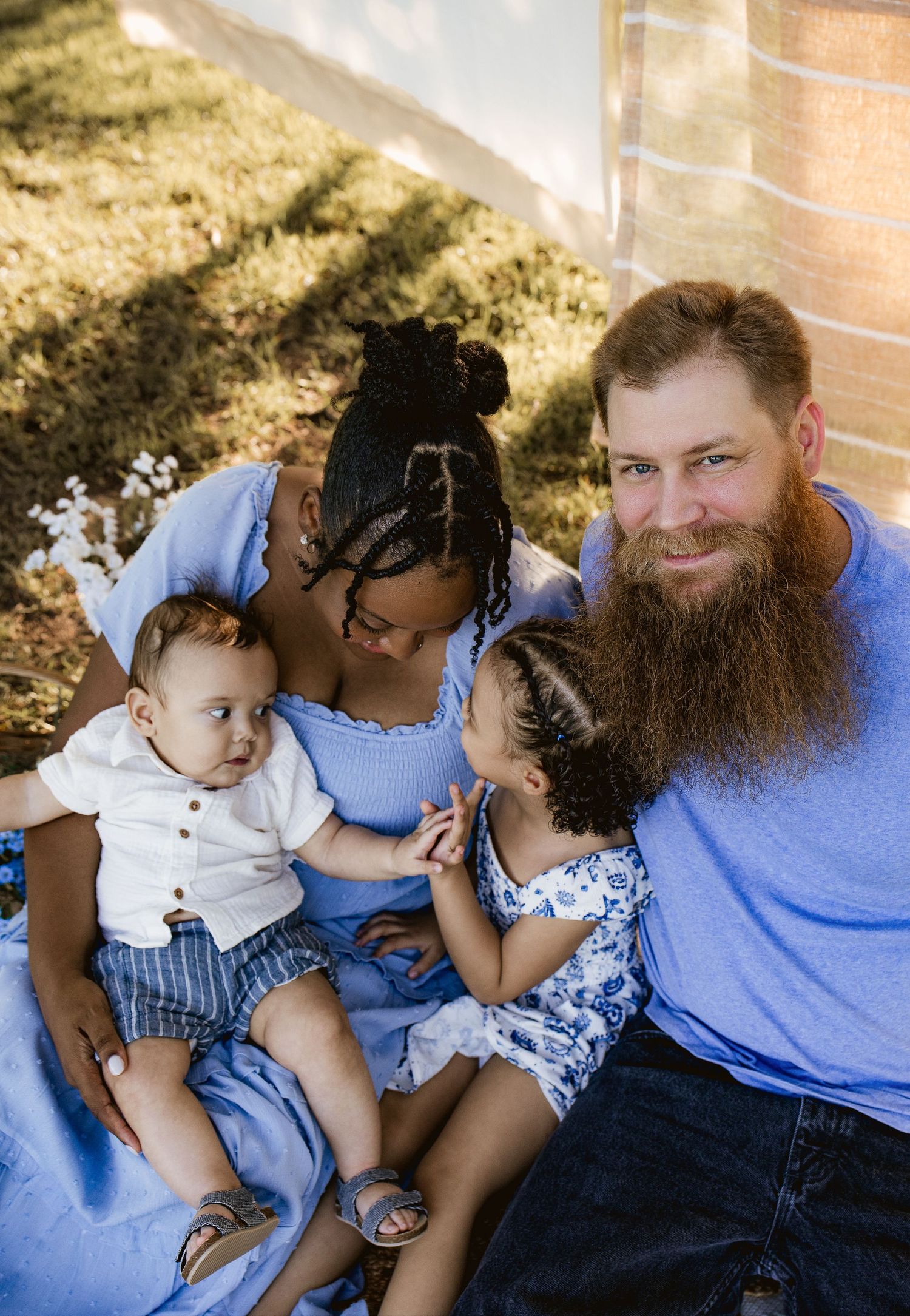 Family of four posing outdoors, parents with two young children in coordinating blue outfits on a sunny day.