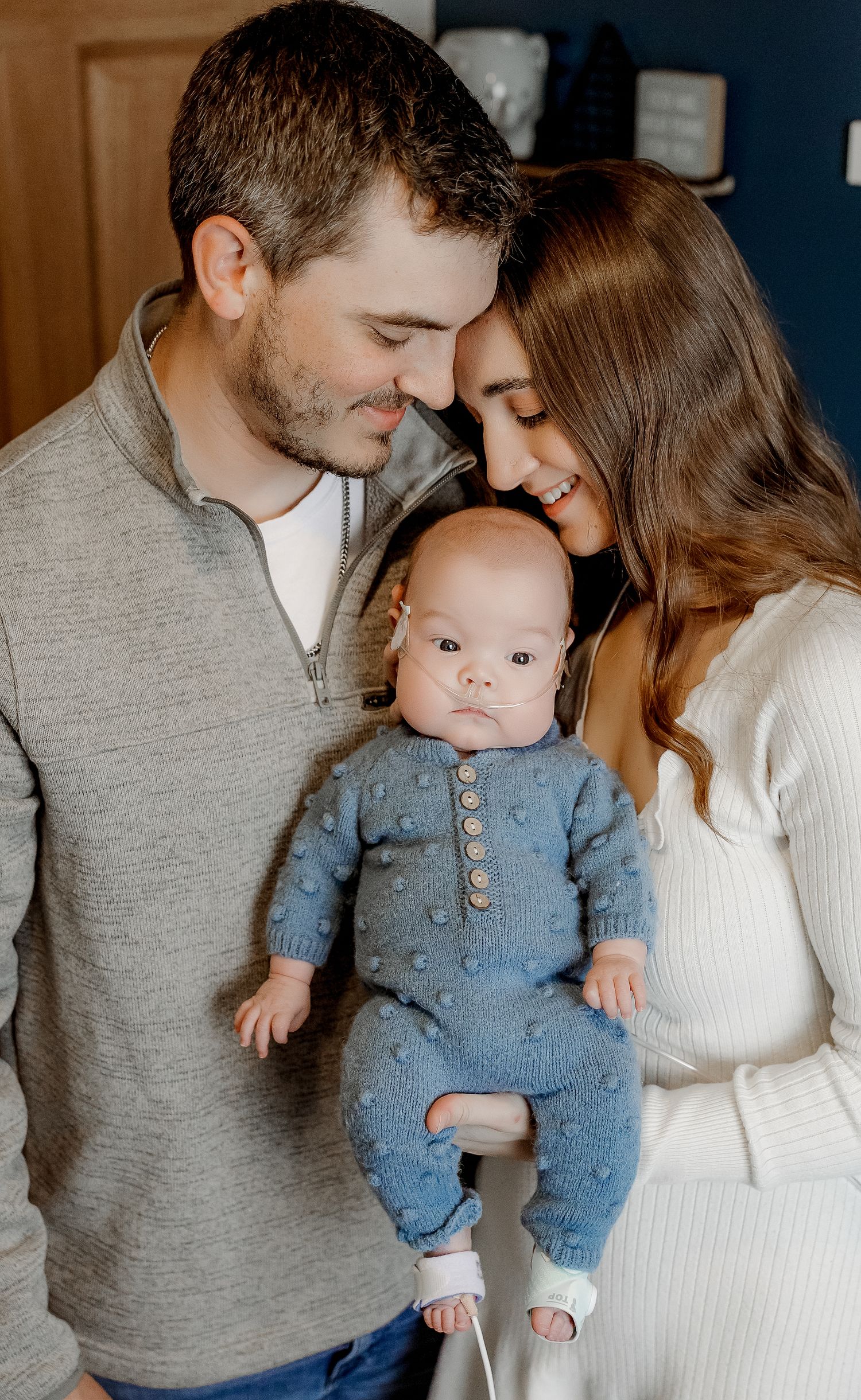 A couple tenderly holds their newborn baby dressed in a blue knit romper, sharing a sweet family moment.