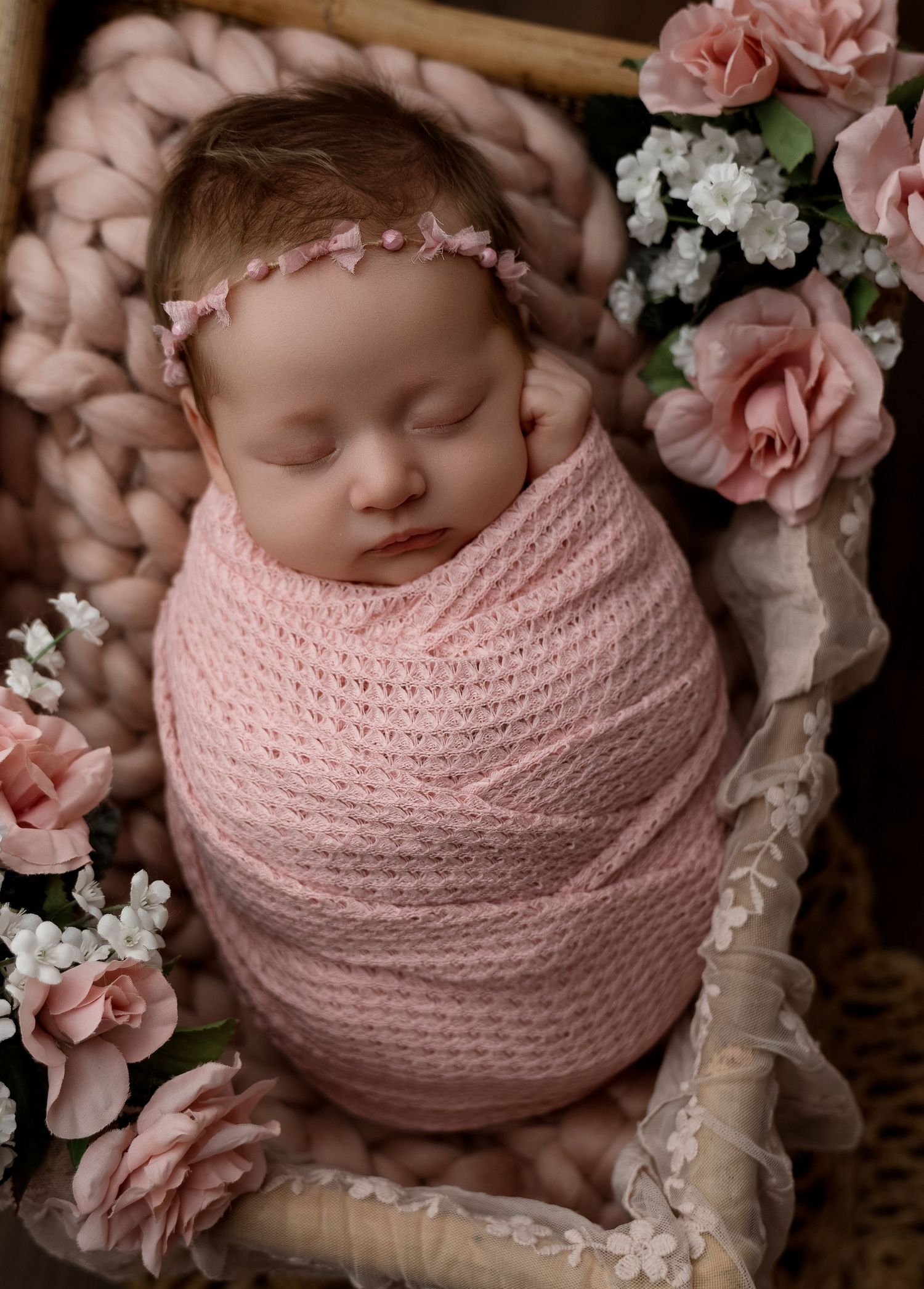 Newborn baby wrapped in pink knit blanket surrounded by roses in a wicker basket.
